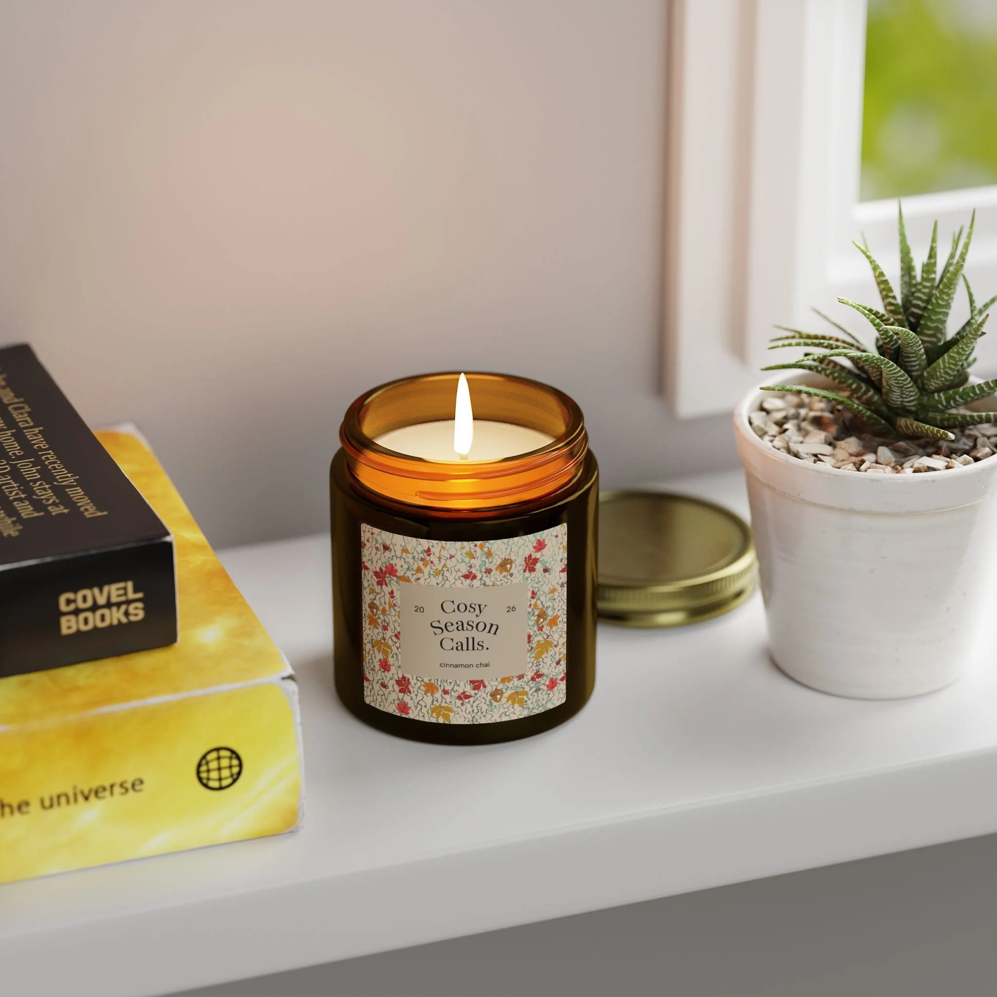 Lit Cosy Season Calls candle on a windowsill next to books and a small plant