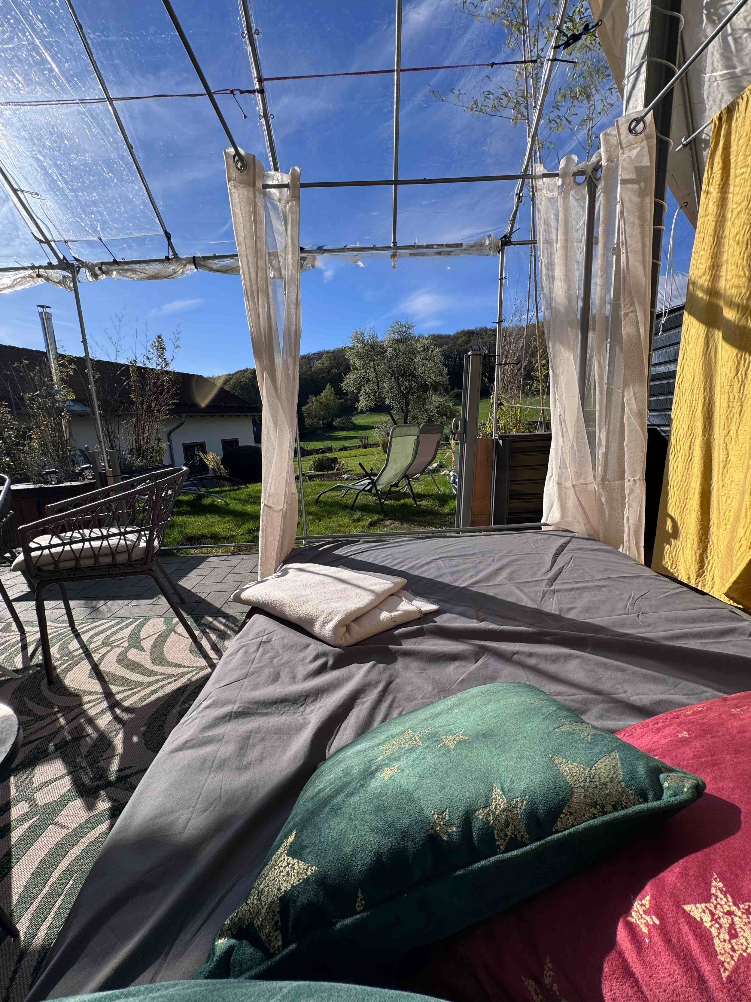 Blick von einem Schlafzimmer auf eine Terrasse mit Stühlen, Liegestühlen, einem Garten mit Bäumen und einem blauen Himmel mit Wolken.