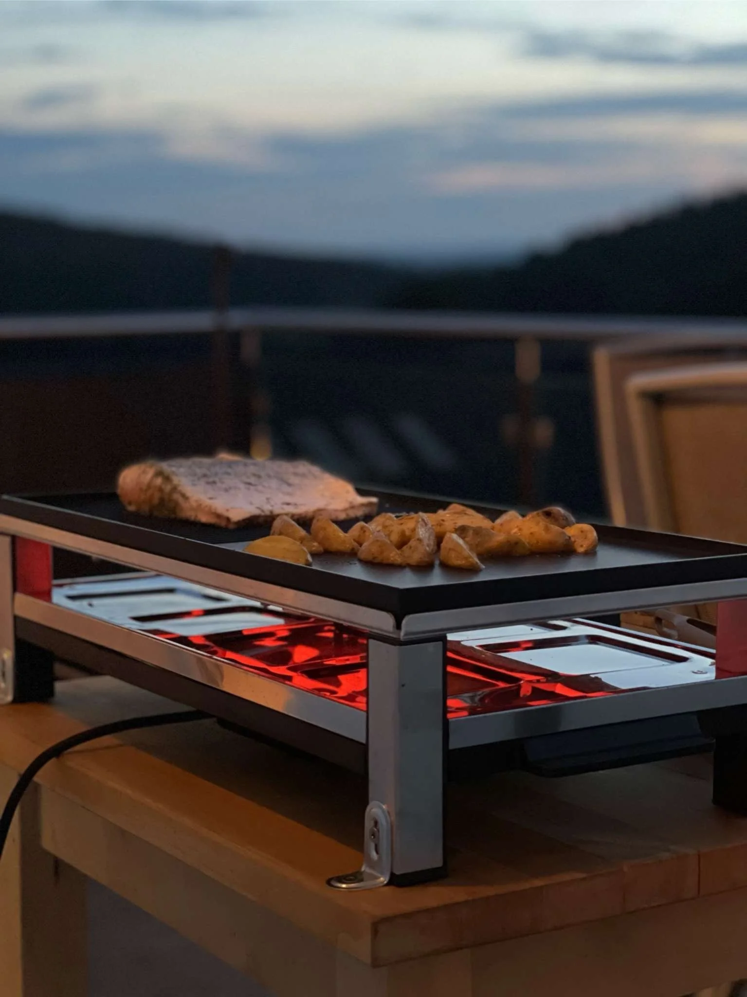 Auf einem elektrischen Grill werden Fleisch und Kartoffeln gegrillt, im Hintergrund ist eine Meereslandschaft sichtbar.