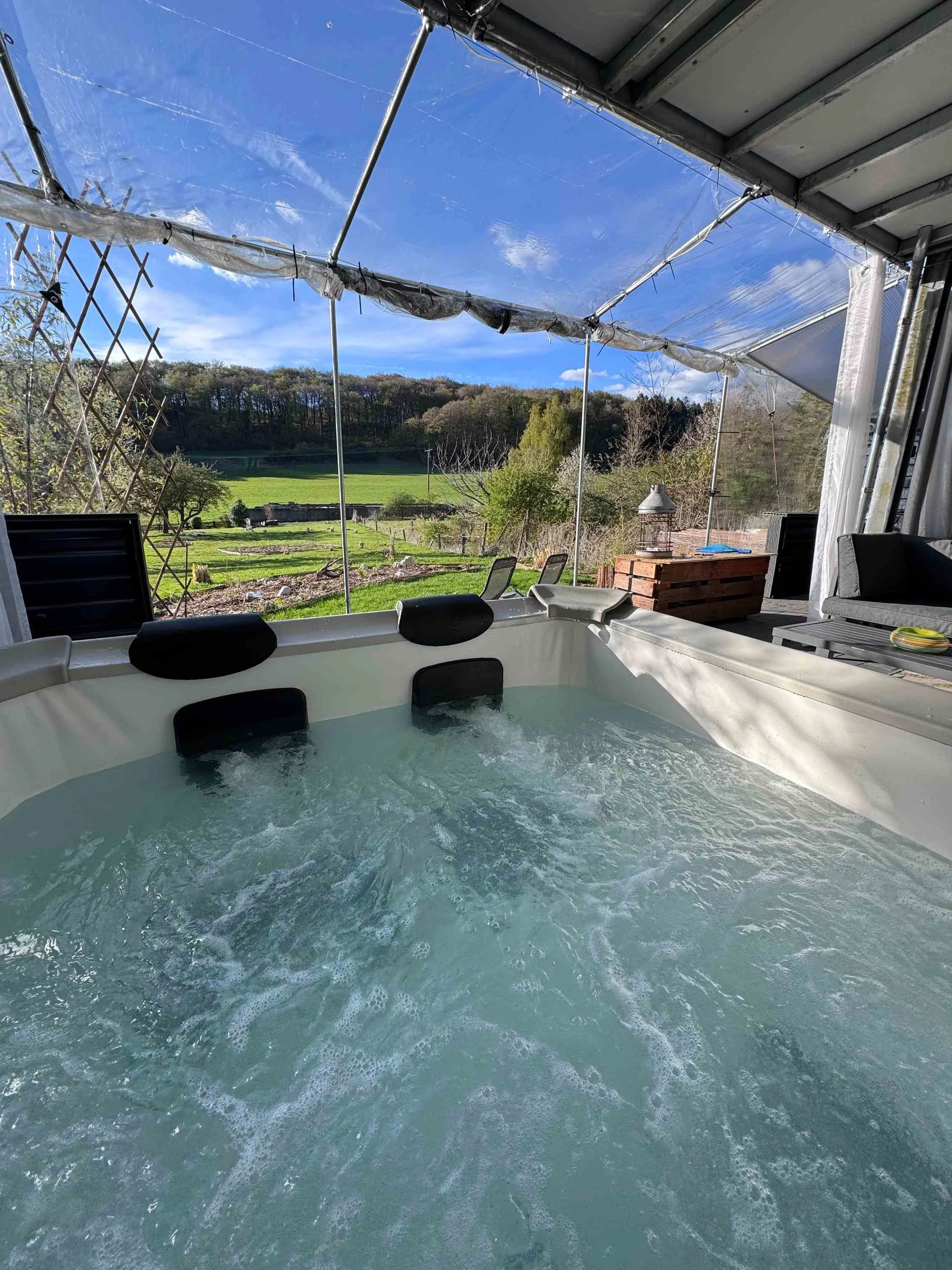 Ein Whirlpool auf einer Terrasse mit Blick auf grüne Felder und Bäume, der unter einem offenen, transparenten Dach steht.