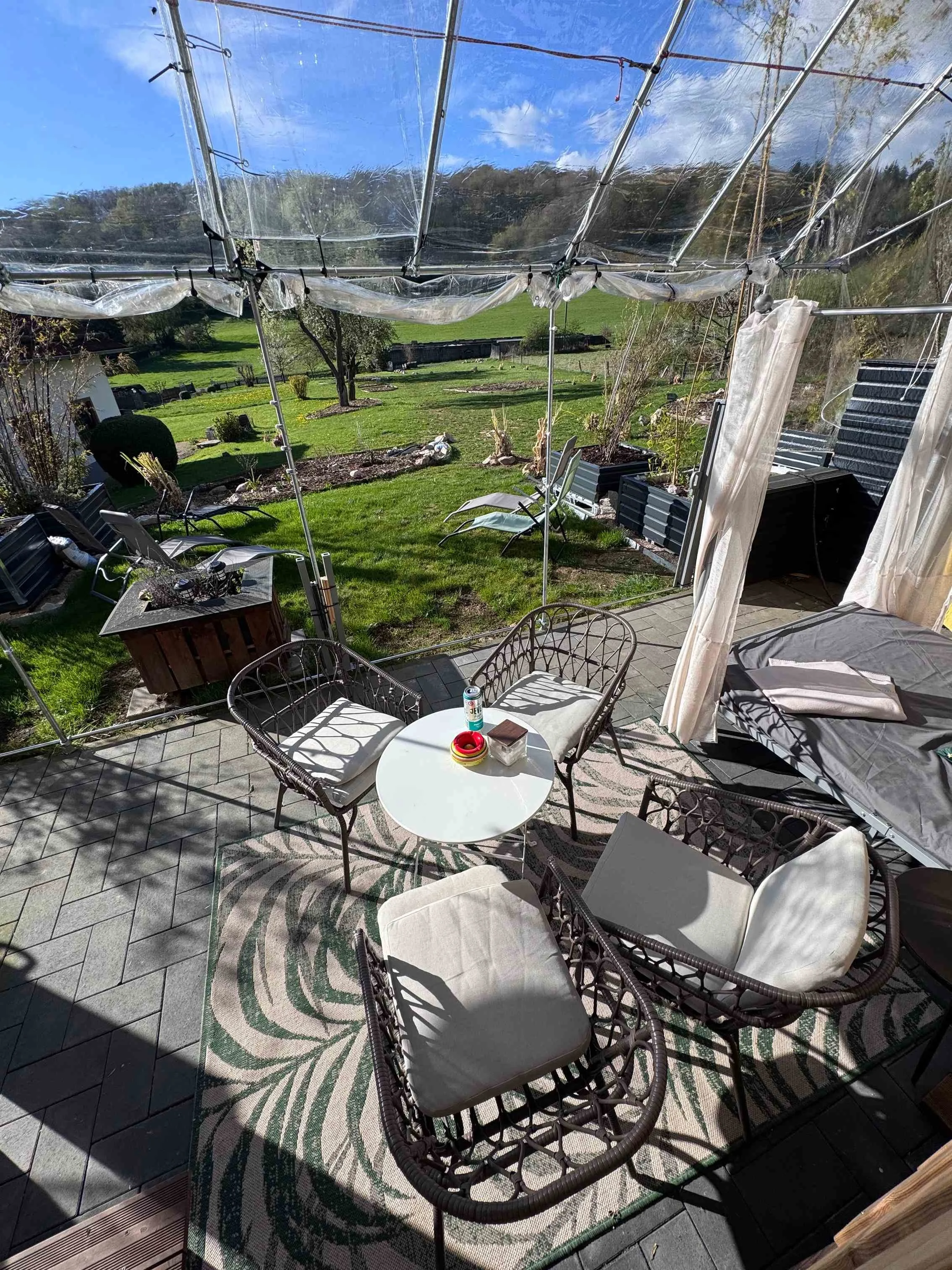 Gemütliche Terrasse mit Gartenmöbeln, Schatten und Blick auf den grünen Garten mit Bäumen und Himmel.