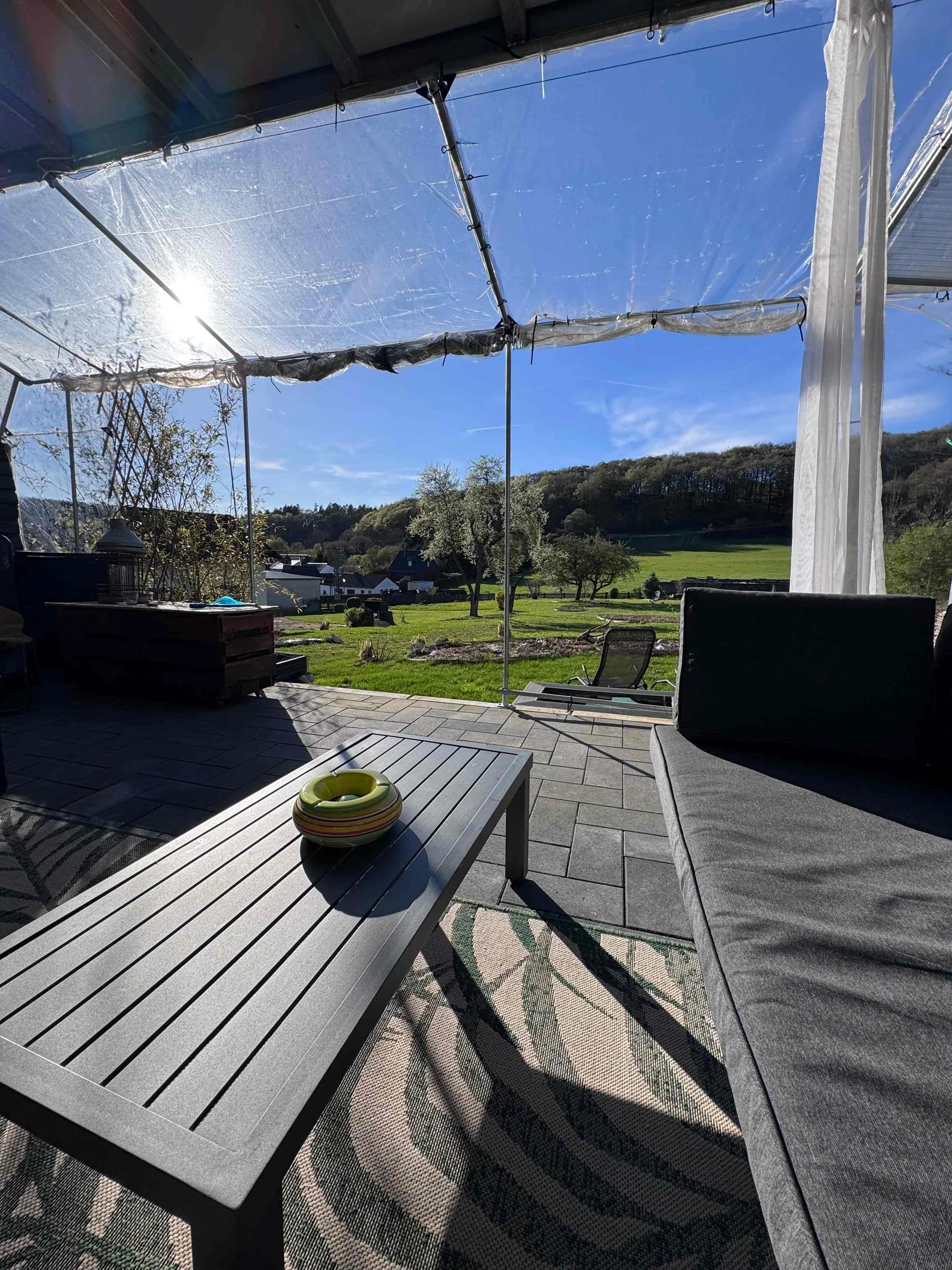 Blick von einer Terrasse mit Sofa, Tisch und Blick auf eine grüne Wiese mit Bäumen und Hügeln bei sonnigem Himmel.