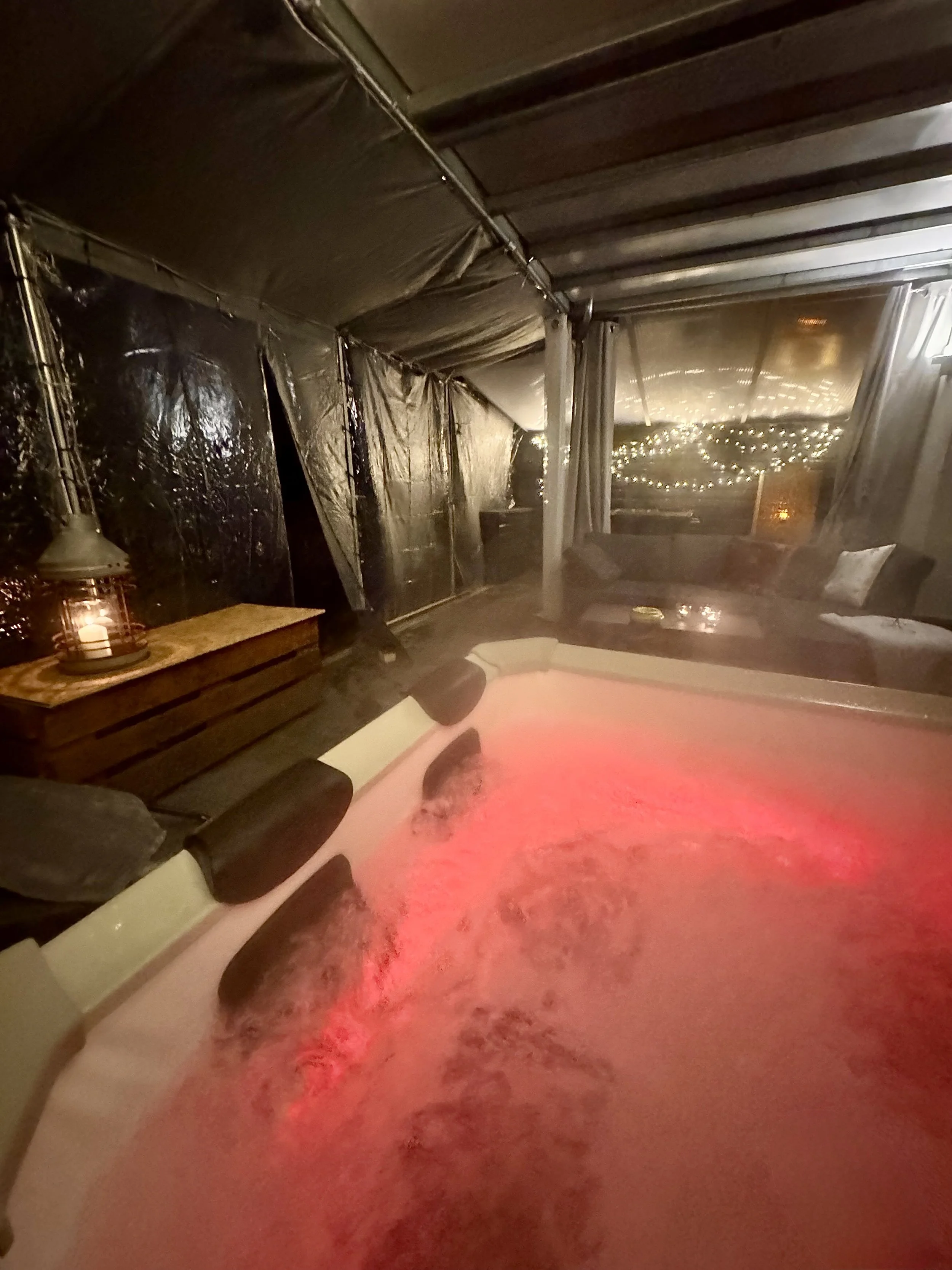Ein Whirlpool mit rot leuchtendem Wasser in einer geschlossenen Terrasse mit schwarzer Folie und Beleuchtung im Hintergrund.