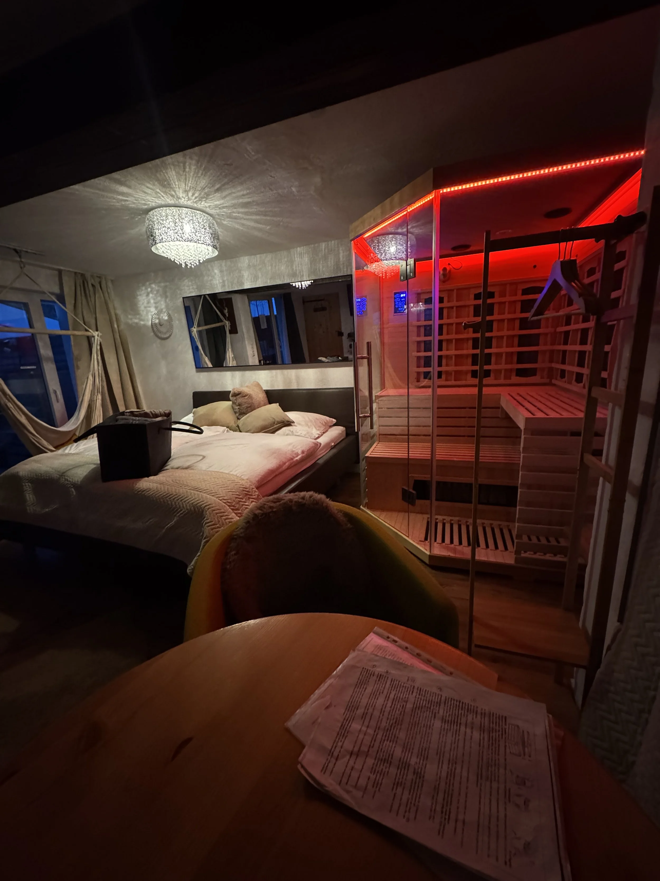 Ein Schlafzimmer mit einem Bett, einer Sauna mit rotem Licht, einem runden Tisch mit Papieren, einem Stuhl mit einem Fell, Gefängnisschrank, einem Spiegel und Vorhängen bei Nacht.