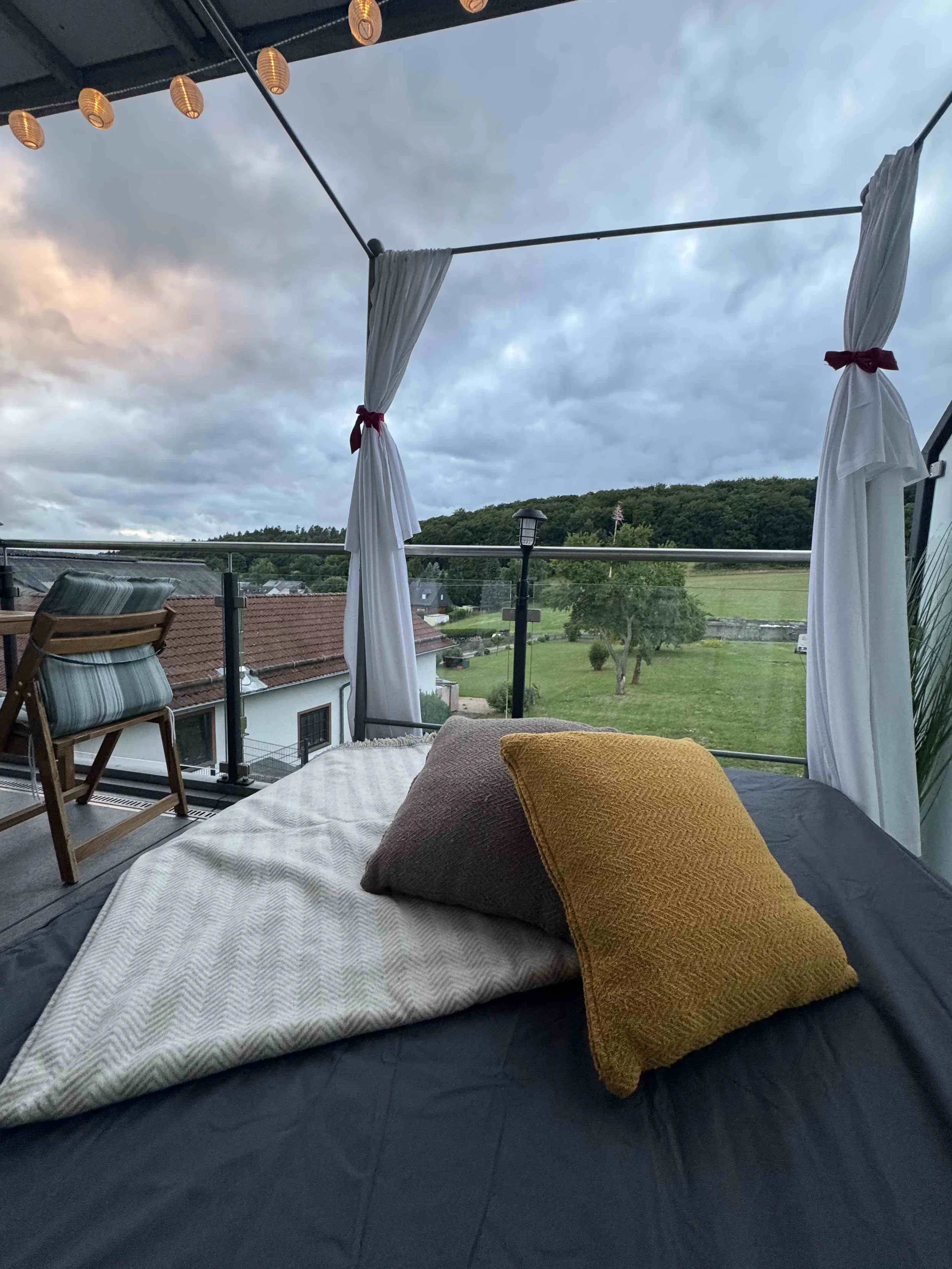 Blick von einem Balkon mit Day Bett und Kissen, weißen Vorhängen, beleuchteten Laternen an der Decke, Blick auf einen Garten mit Bäumen und Hügel im Hintergrund, bewölkter Himmel.
