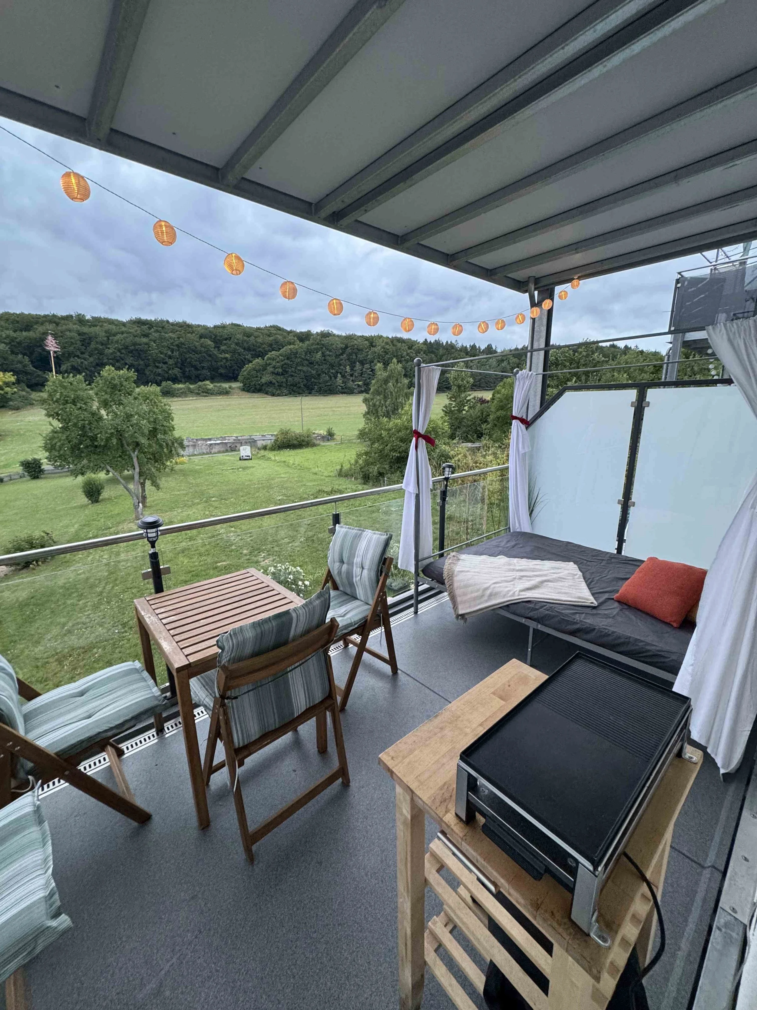 Balkon mit Sitzgelegenheiten, Daybed, Tisch, Grill, mit Blick auf grüne Wiese und Bäume, abgedeckt mit Lichterkette und Vorhängen