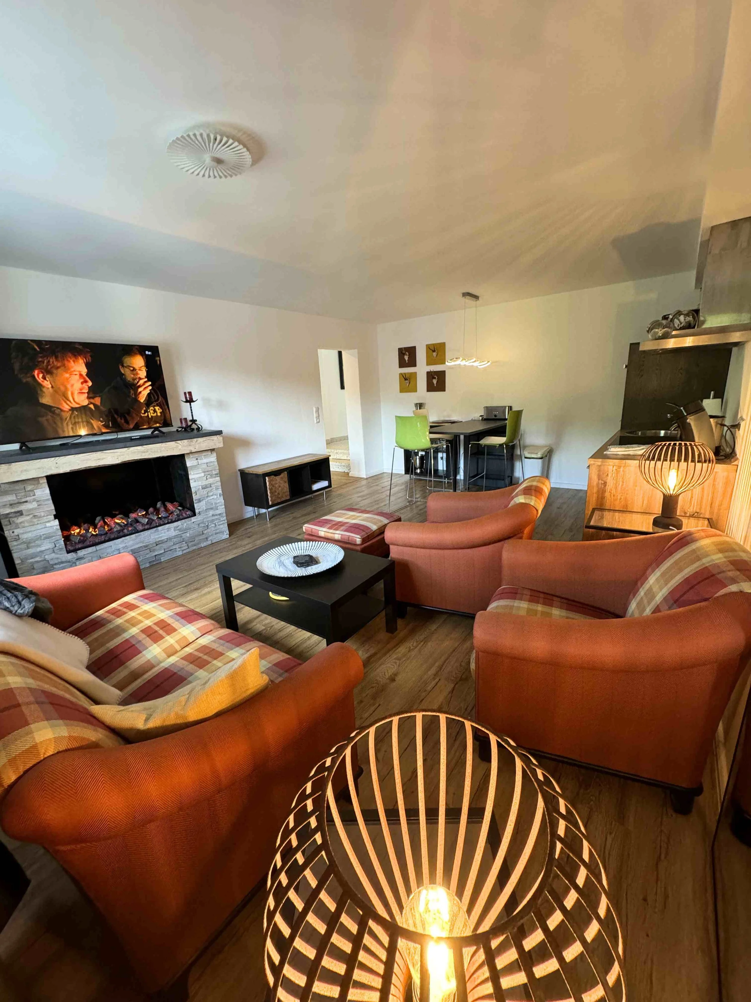 Gemütliches Wohnzimmer mit vier roten Polstermöbeln, einem schwarzen Couchtisch, einem Kamin mit Fernseher, einem Essbereich im Hintergrund, einem modernen Lampenschirm und einem holzvertäfelten Sideboard.