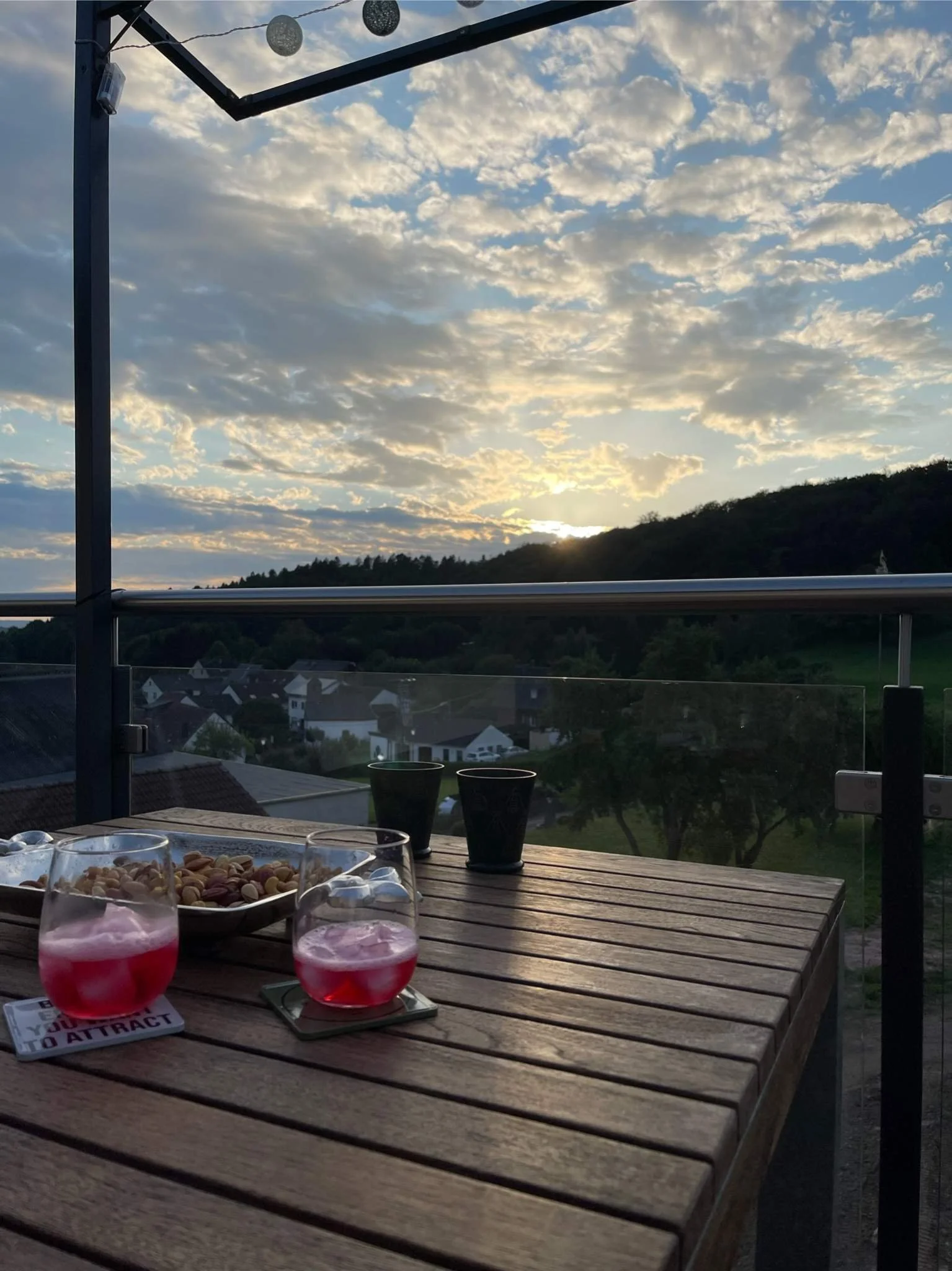 Ein Balkon mit einem Holztisch, auf dem zwei Gläser mit rosa Getränken und eine Schale mit Nüssen stehen. Es gibt auch zwei leere schwarze Töpfe. Im Hintergrund sieht man eine Landschaft mit Hügeln, Bäumen und einem bewölkten Himmel bei Sonnenunterga