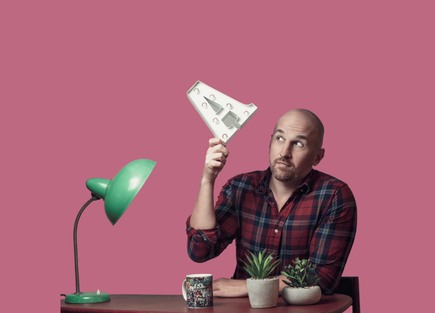 Andy Selway - Local Bicester based Hypnotherapist, supporting local small business owners - coaching and therapy, sat at desk, pink backdrop, in a flannel read shirt, holding up an ornament with lights of an "A" - signifying business success