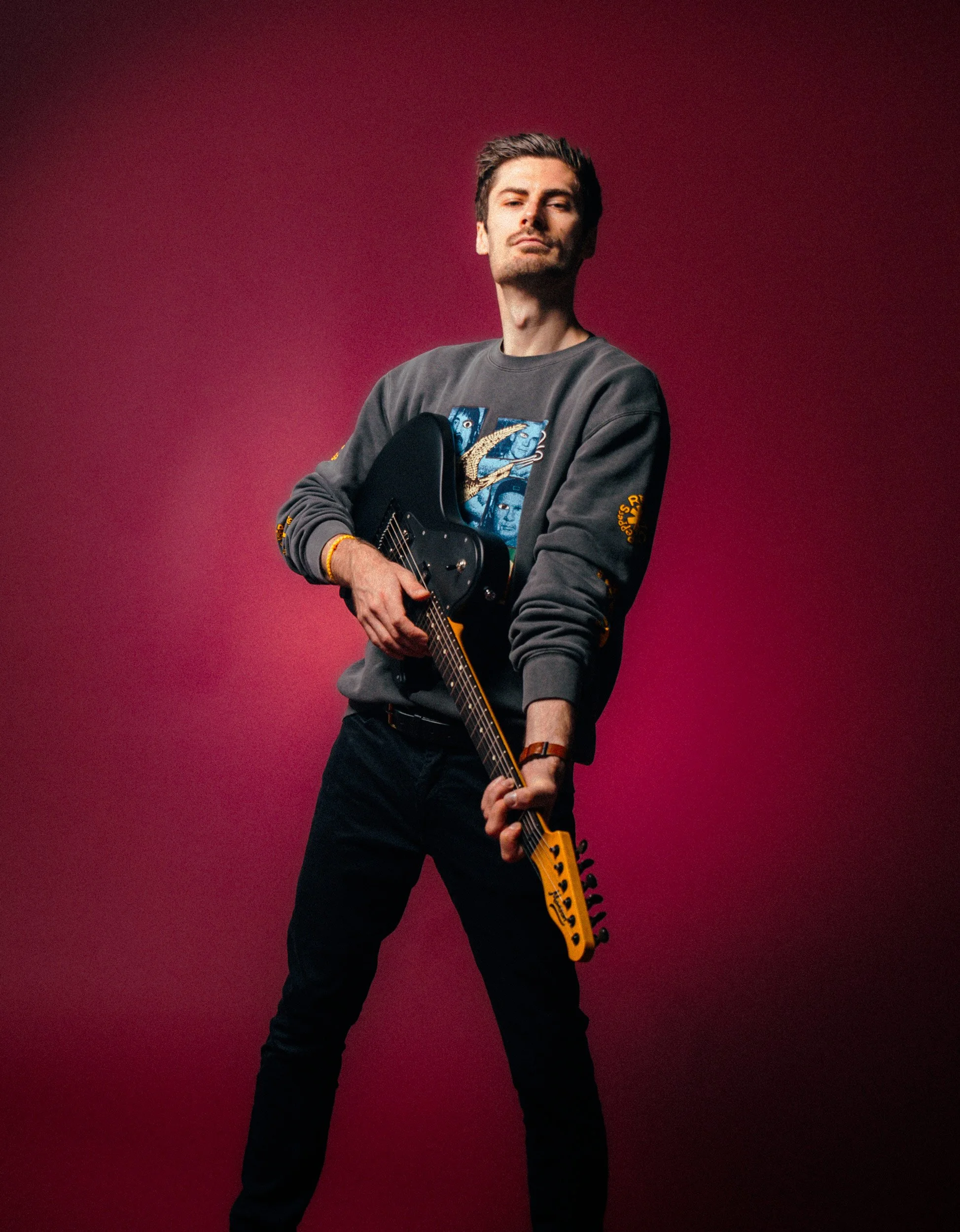 A man with dark hair and light skin, posing with a black electric guitar against a red background. He is wearing a gray sweatshirt with colorful designs on the front and yellow text on the sleeve, and black pants.