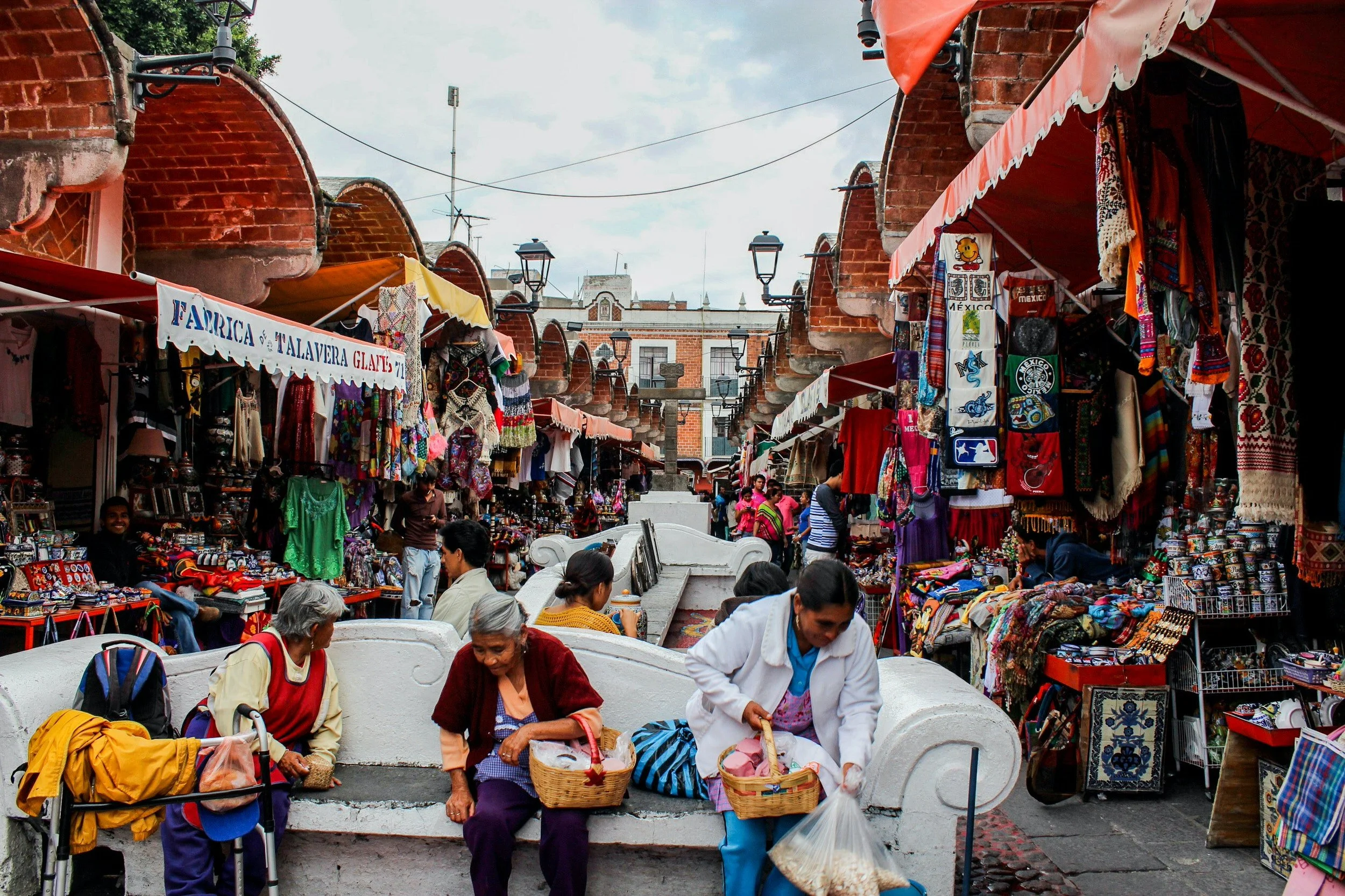 Market with stalls selling textiles, clothing, and souvenirs, people sitting on a white bench, and vendors attending to customers, with brick buildings and street lamps in the background.