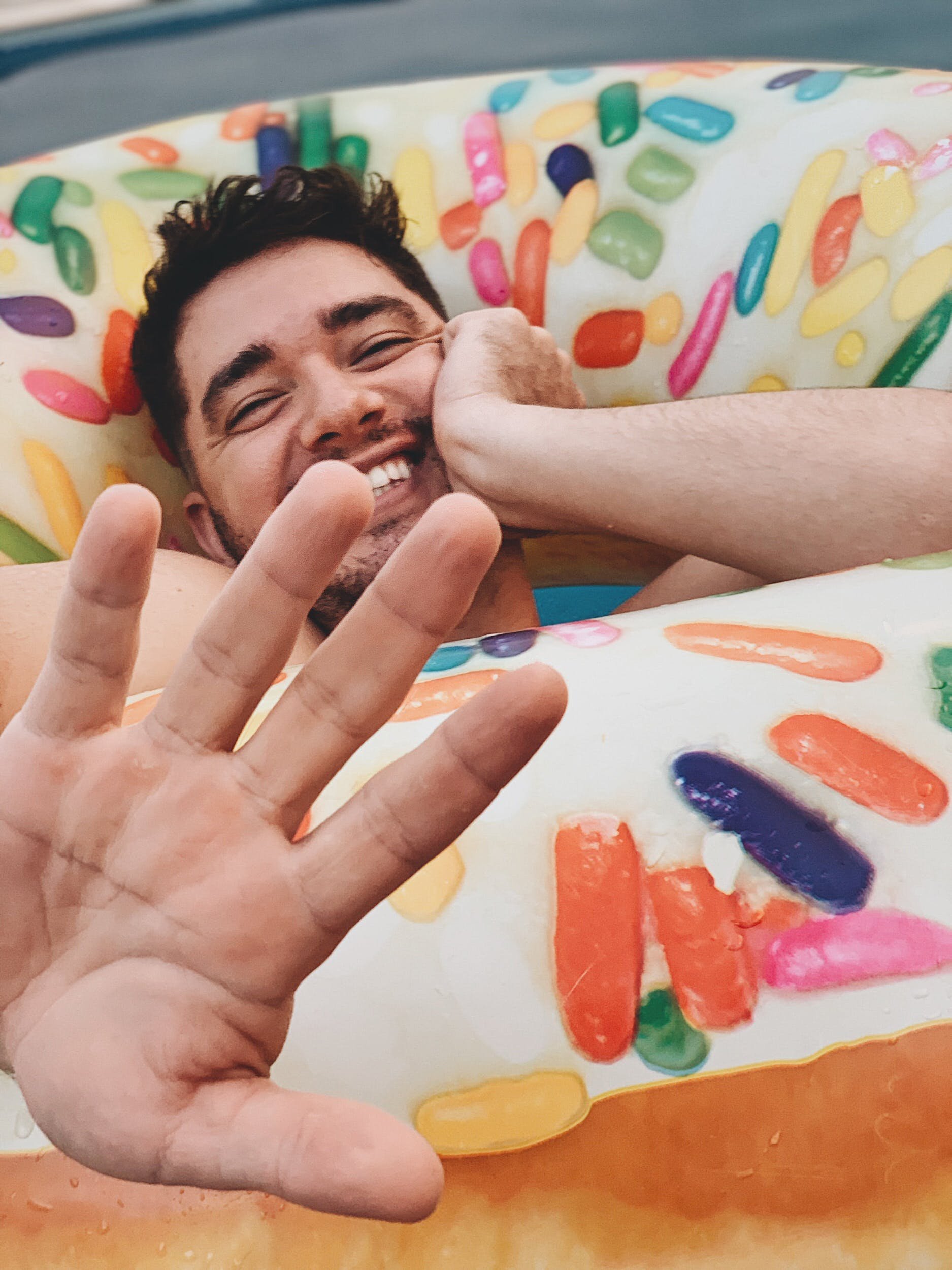 A man smiling and lying on a float decorated like a giant gummy candy, with a hand showing three fingers in the foreground.