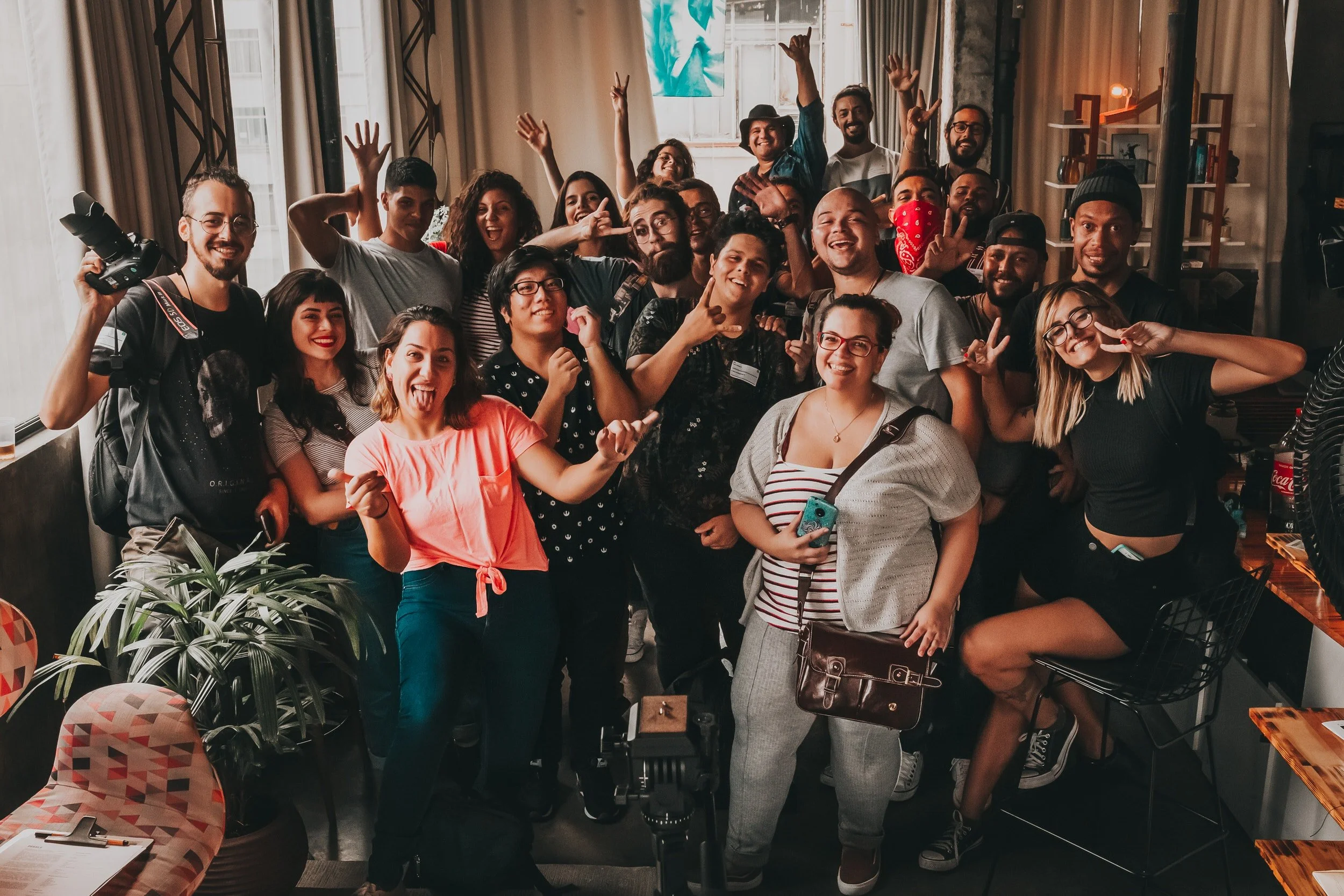 Group of diverse people smiling and posing for a photo indoors, some making peace signs and gestures, with a camera and photography equipment visible.