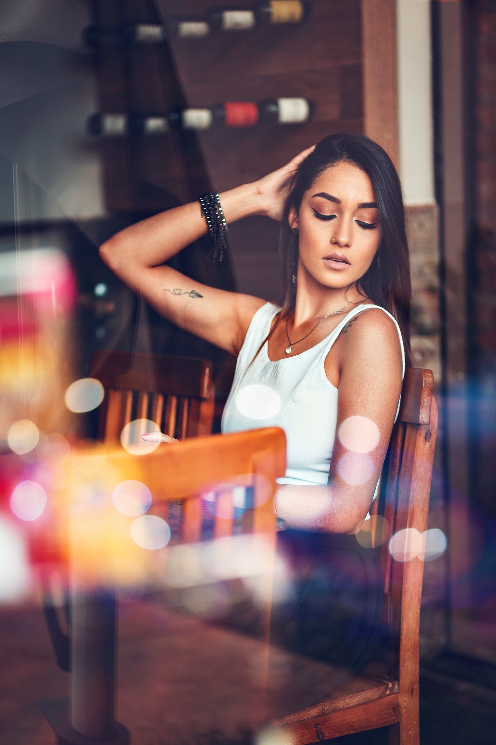 A young woman with dark hair and makeup sitting in a cozy cafe, wearing a white tank top and jewelry, captured through a window with colorful bokeh reflections.