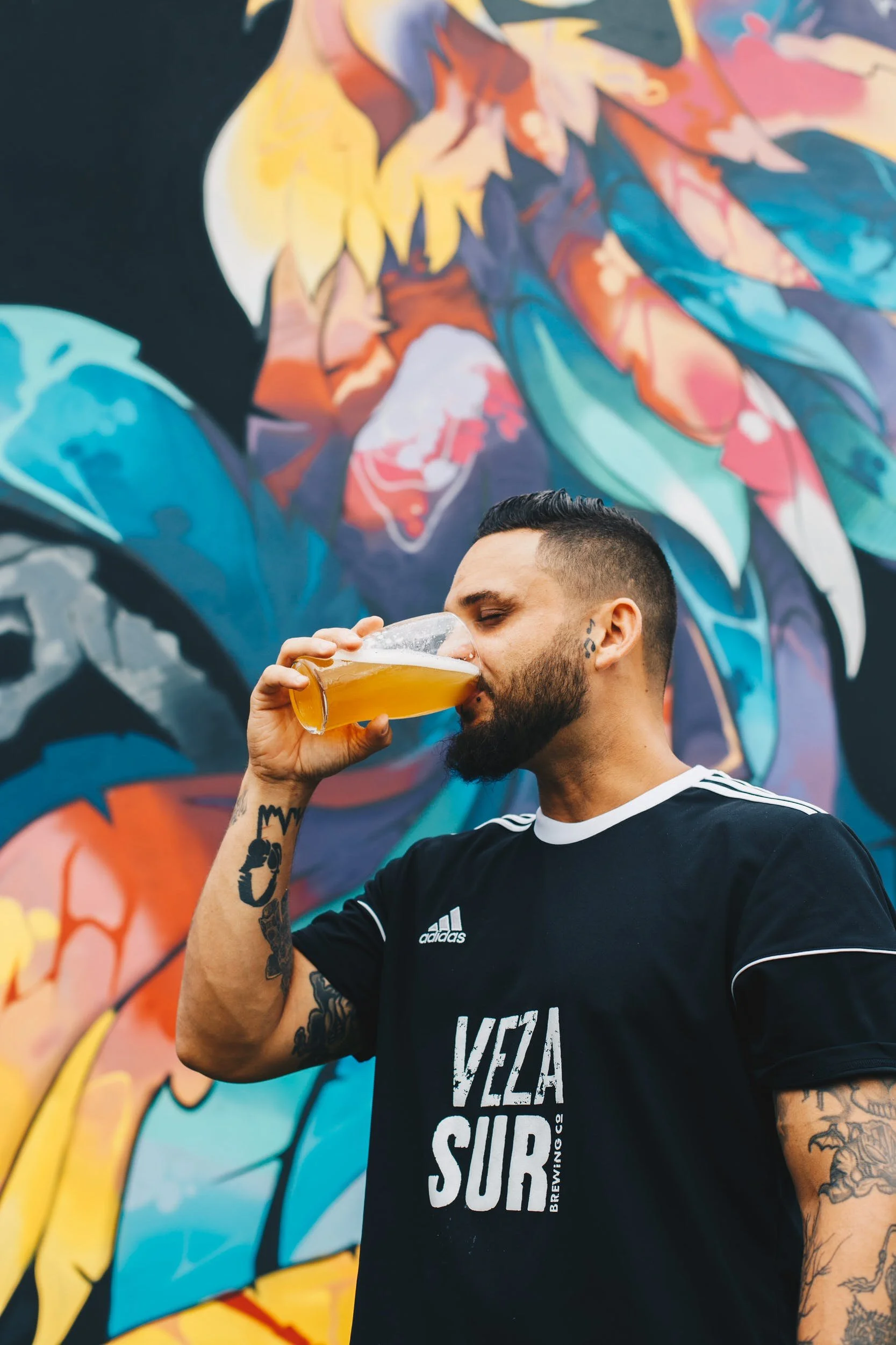 A man with tattoos and a beard drinks beer from a glass in front of a colorful mural.