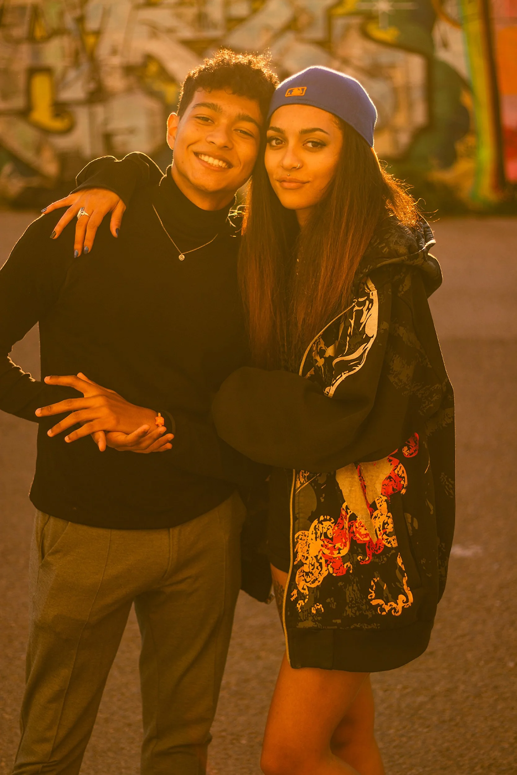 A young man and woman standing close together outdoors in front of a colorful graffiti wall, smiling at the camera during sunset or golden hour. The man has short curly hair, is wearing a black turtleneck, gray pants, and a silver necklace. The woman