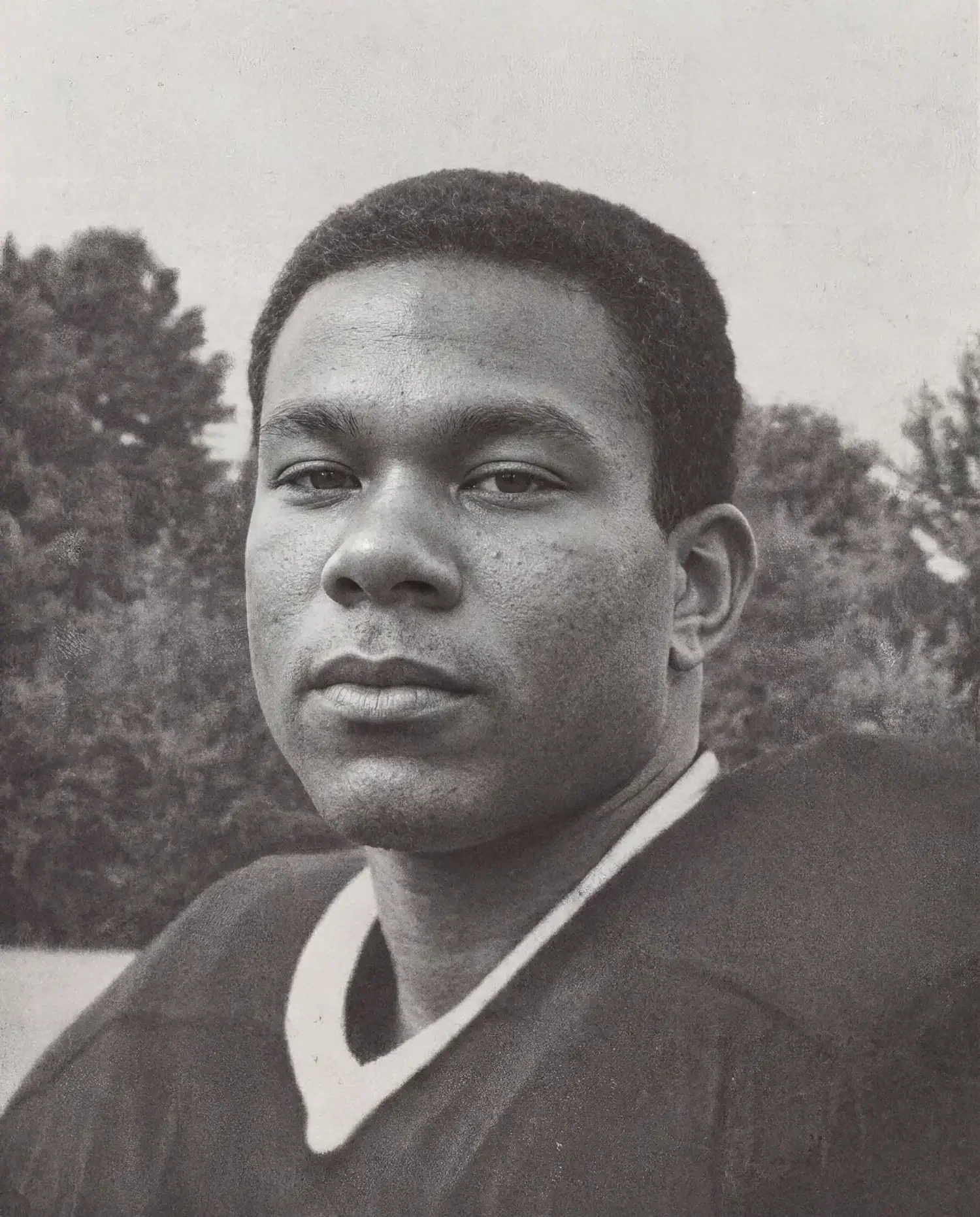 Black and white photograph of a young man with short hair, wearing a sports jersey, outdoors with trees in the background.