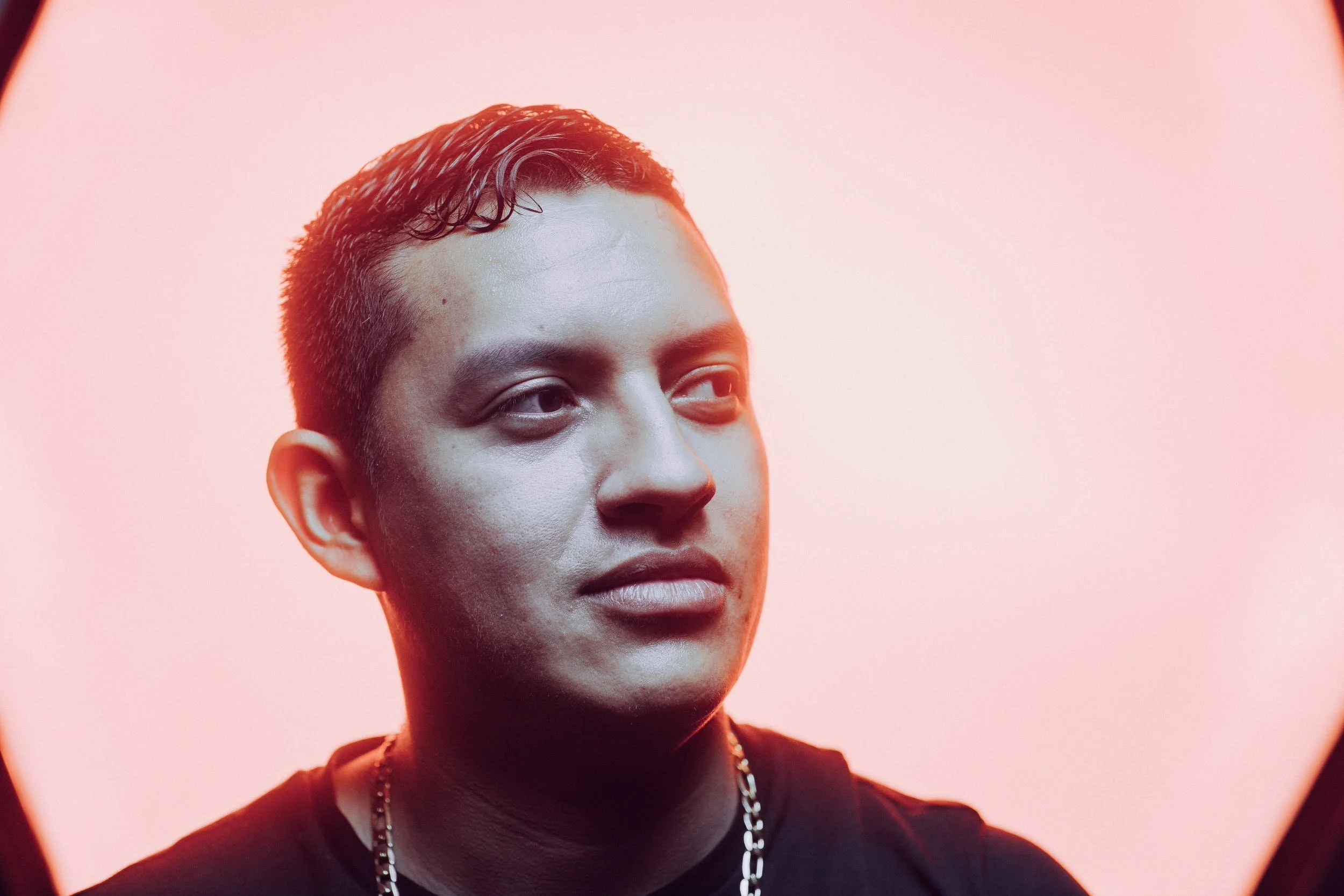Close-up of a young man with short dark hair, wearing a black shirt and chain necklace, against a pink background.