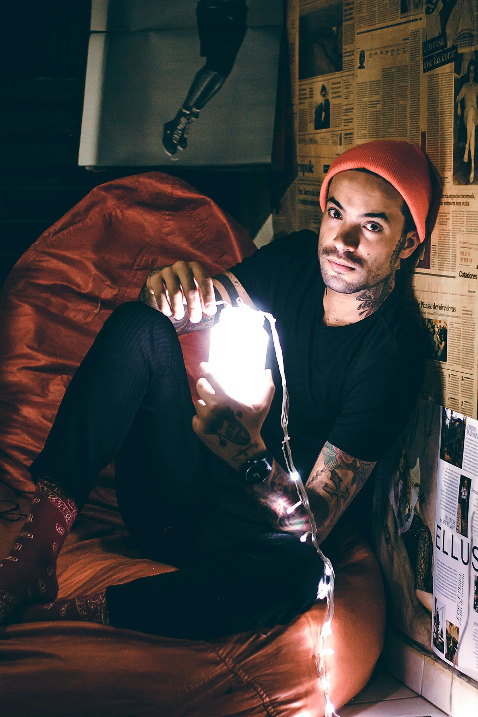 A young man with tattoos and wearing a red beanie sitting on a bed in a dimly lit room, holding a glowing smartphone, with postcards and posters on the wall behind him.