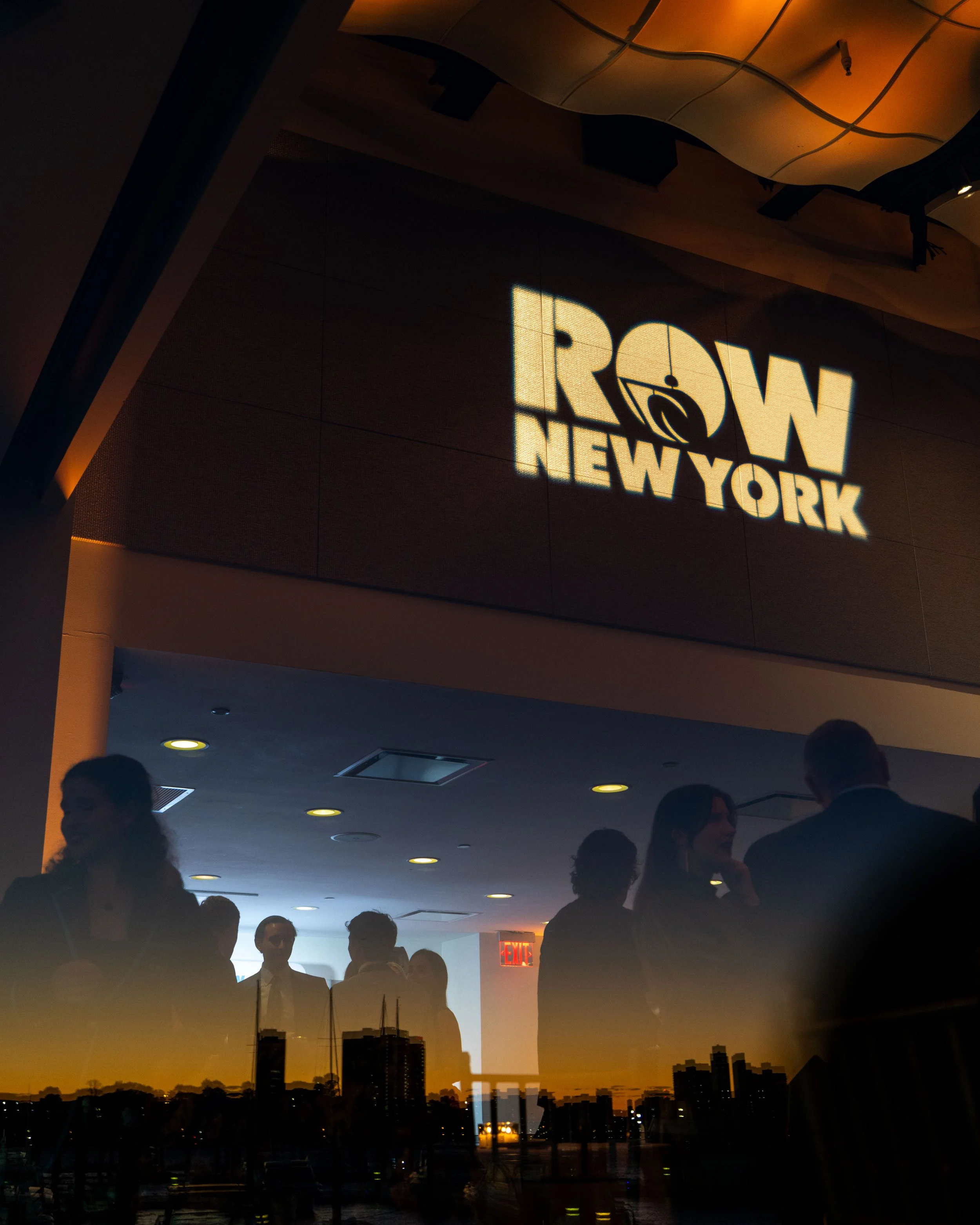 A projection on a wall for Row New York, along with silhouettes of people in a dark indoor setting, and a city skyline reflection at the bottom at a fundraising gala produced by JKM Events.