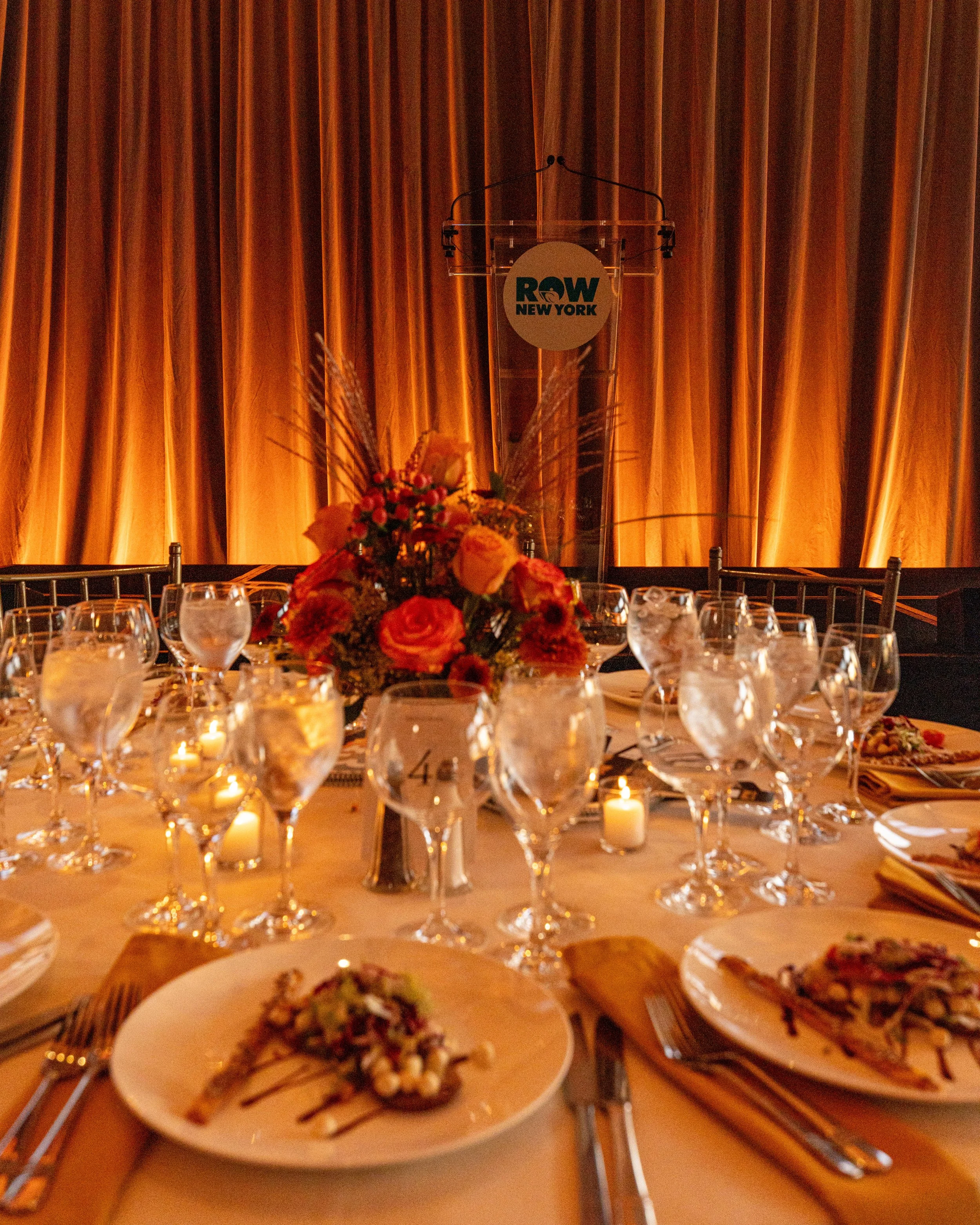 Elegant banquet table set with multiple wine glasses, plates of food, lit candles, and floral centerpiece in front of a gold curtain backdrop at a fundraising gala for Row New York, produced by JKM Events.