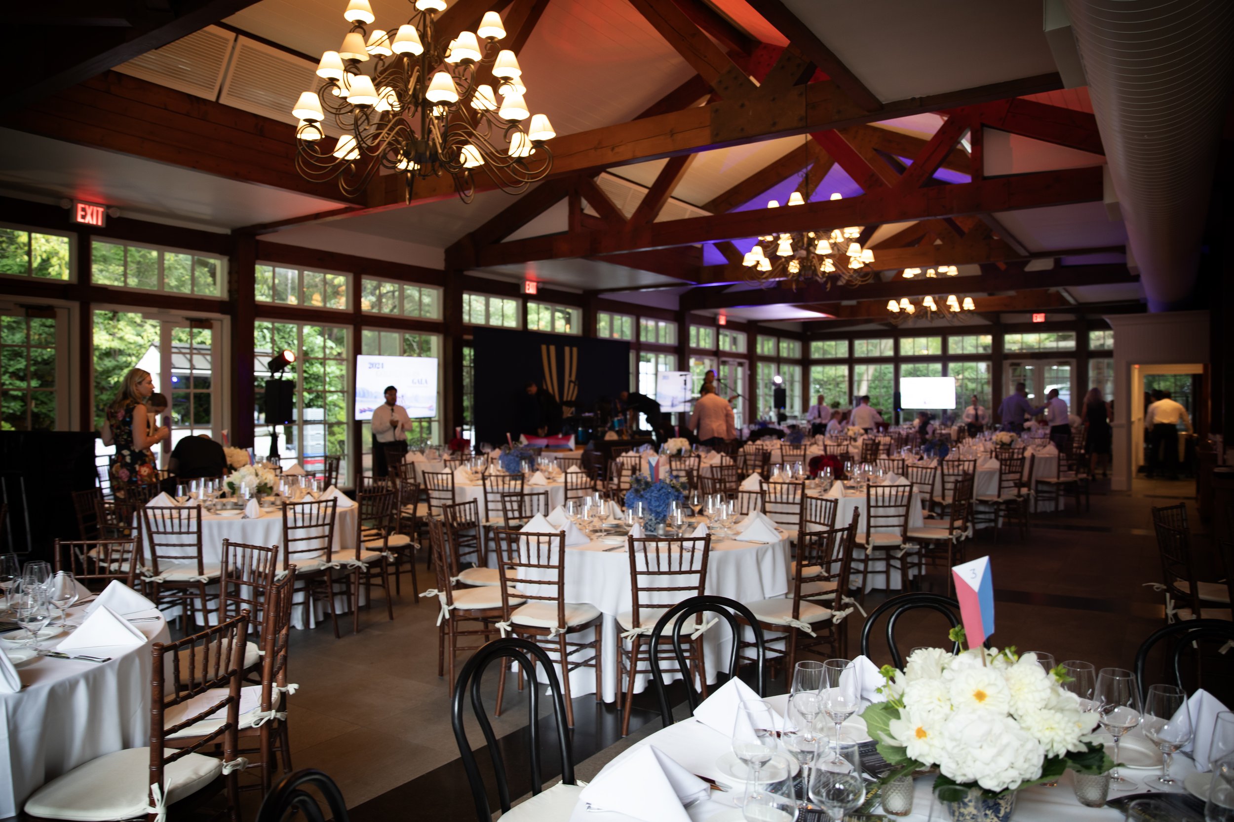 Fundraising gala decorated with white and blue floral centerpieces, set with glassware and napkins, and surrounded by wooden chairs with white cushions, produced by JKM Events.