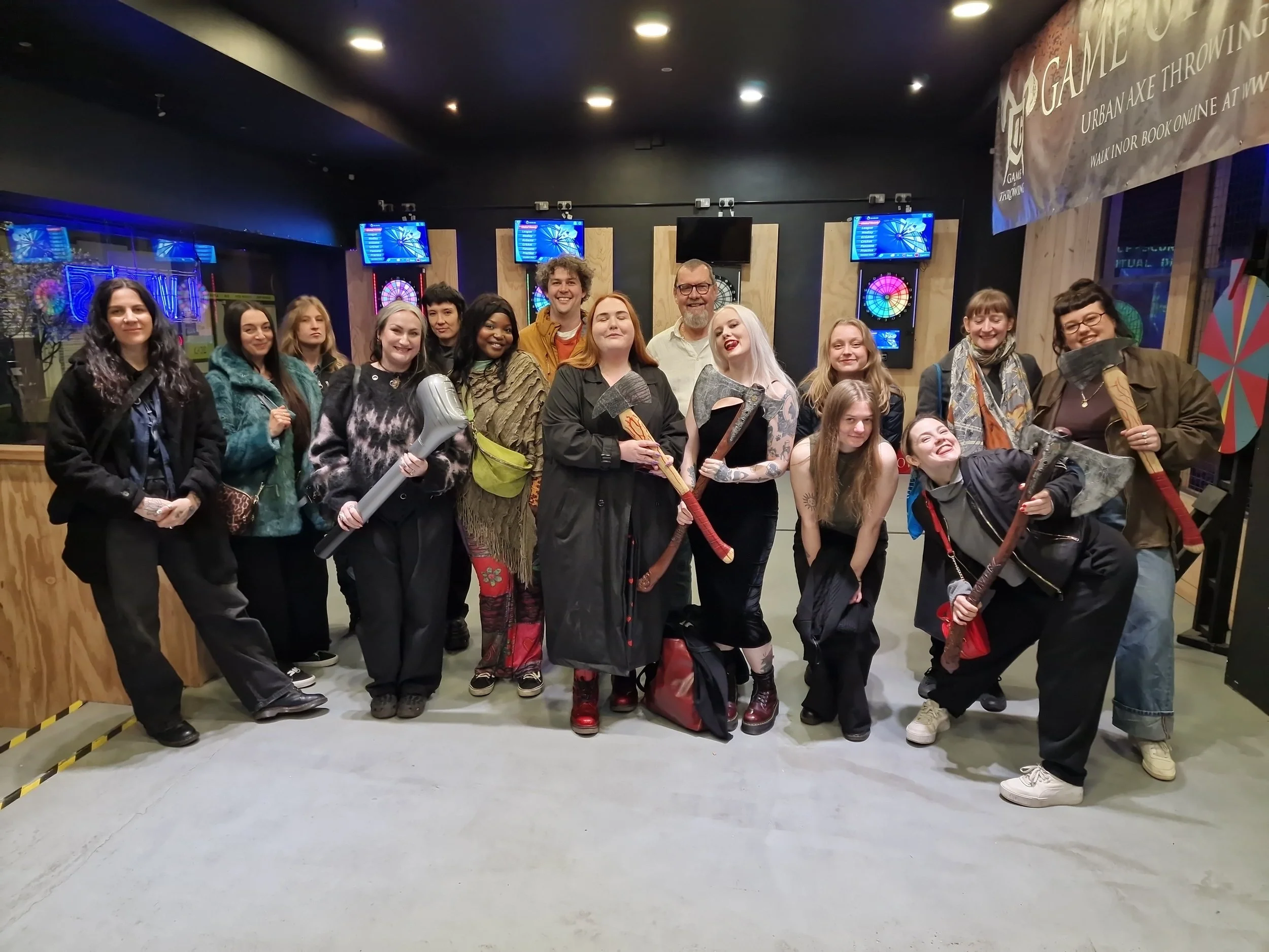 A group of people posing for a photo at an indoor arcade, holding various axes and a large foam axe, with multiple digital dartboards on the wall behind them.