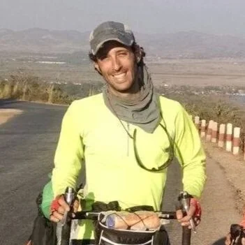 Person wearing a bright yellow shirt and cap, standing outdoors with a bicycle on a road, mountainous landscape in the background.
