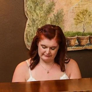 Person with brown hair sitting at a piano, wearing a white top, with a painting of potted trees on the wall behind.