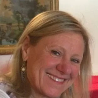 Smiling person with blonde hair, wearing earrings, with a painting in the background.