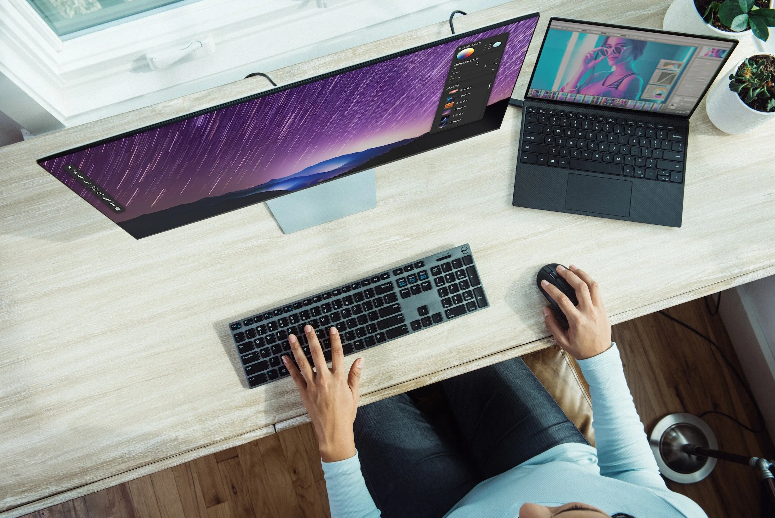 A person using a desktop computer with ultrawide monitor, laptop, keyboard, and mouse on a wooden desk.