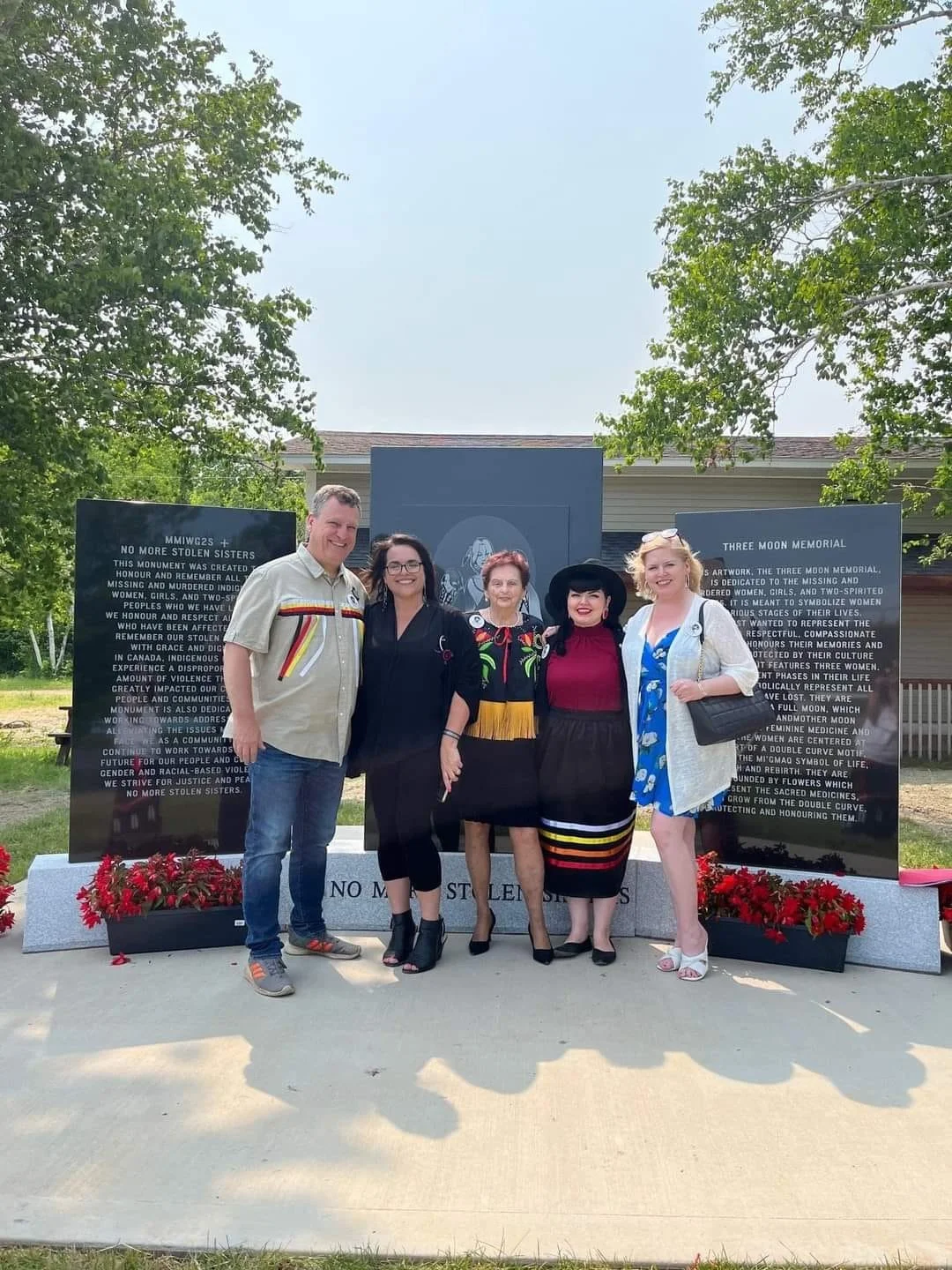 Five women and one man standing together in front of a memorial plaque with red flowers at the base, outdoors on a sunny day with trees in the background. MMIW2+