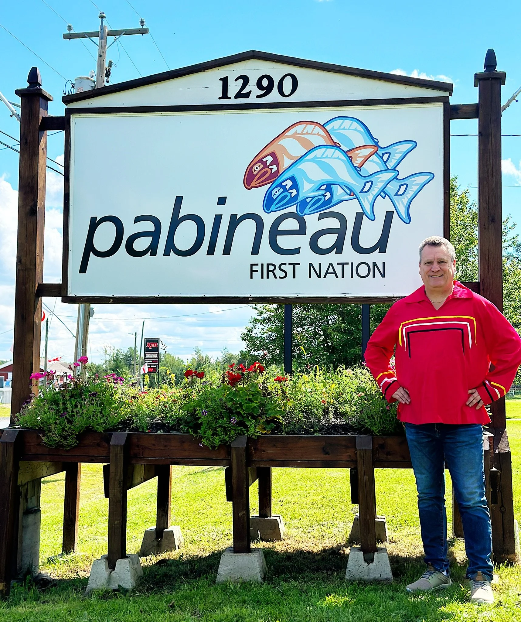 A man in a red shirt standing next to a sign that says "Pabineau First Nation" with a fish logo and the number 1290.