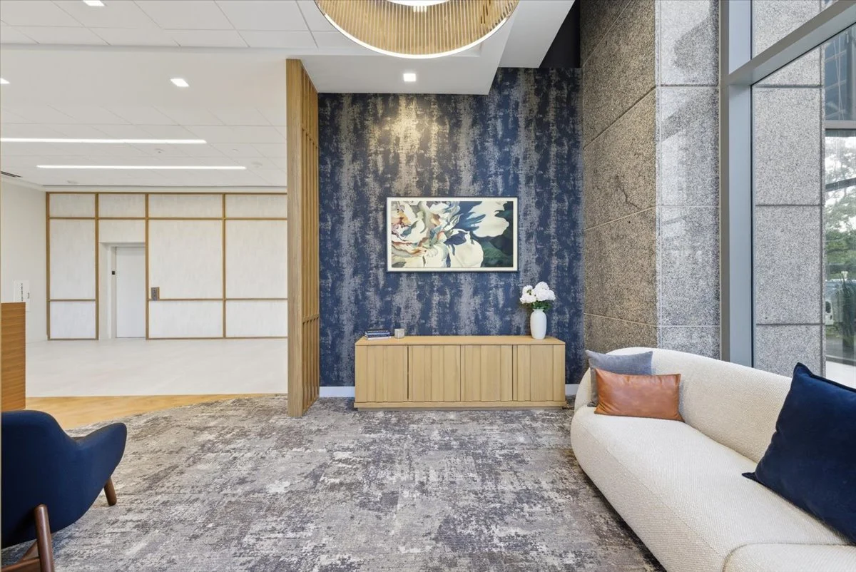 Modern seating area with a white couch, blue and brown pillows, a small wooden cabinet with a flower vase, a blue textured accent wall with framed TV, large windows, and an open space in the background.