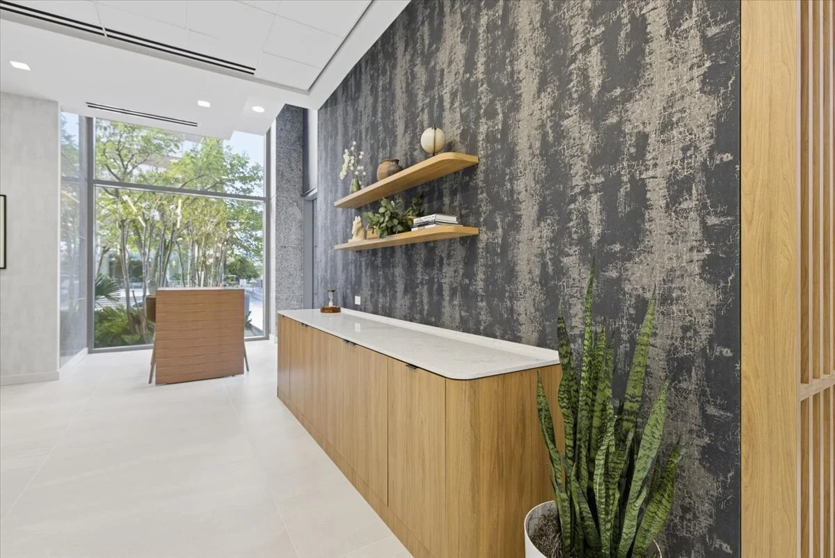Modern interior with a wooden cabinet and floating shelves on a textured black wall, a large potted plant, and a window with trees outside.