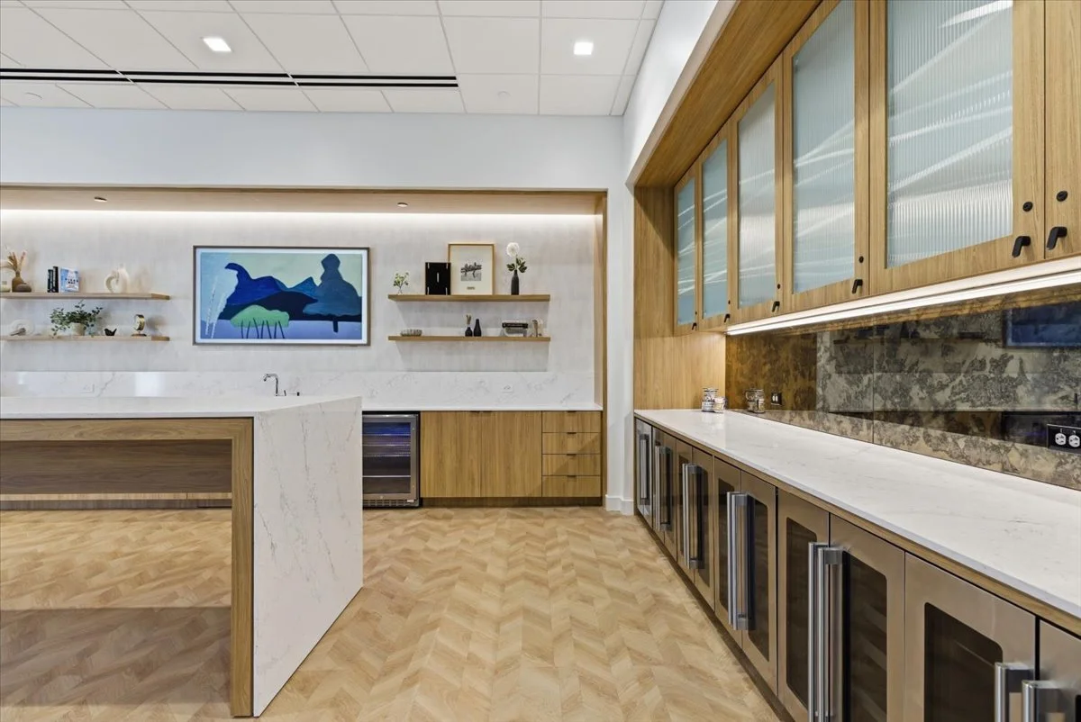 Modern lounge with wooden cabinets, marble countertops, and a glass-fronted wine fridge, featuring wall-mounted shelves with decorative items and a vibrant abstract painting on the wall.