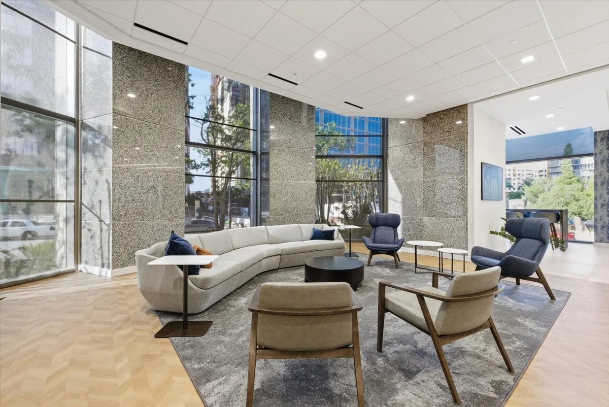 Modern lounge area with large windows, a curved white sofa, four armchairs, and small tables, with a view of trees and buildings outside.