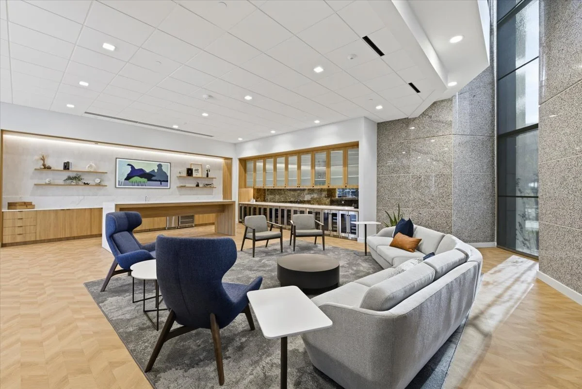 Modern lounge area with sofas, armchairs, and a rug, featuring a kitchenette with wooden cabinets and glass doors, and a large window allowing natural light.