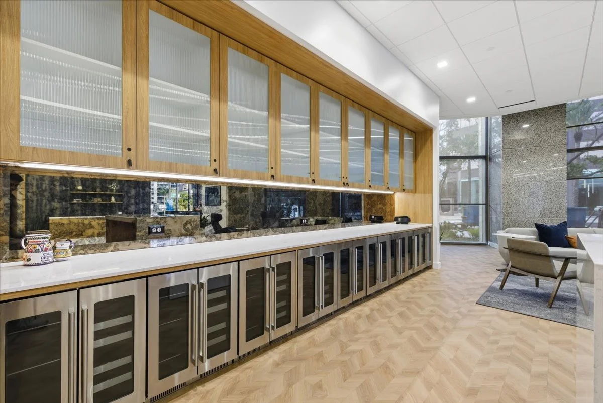 Modern lounge area with a long built-in counter, glass-front wine refrigerators, upper cabinets with frosted glass doors, large windows, seating area with chairs, and light wood flooring.