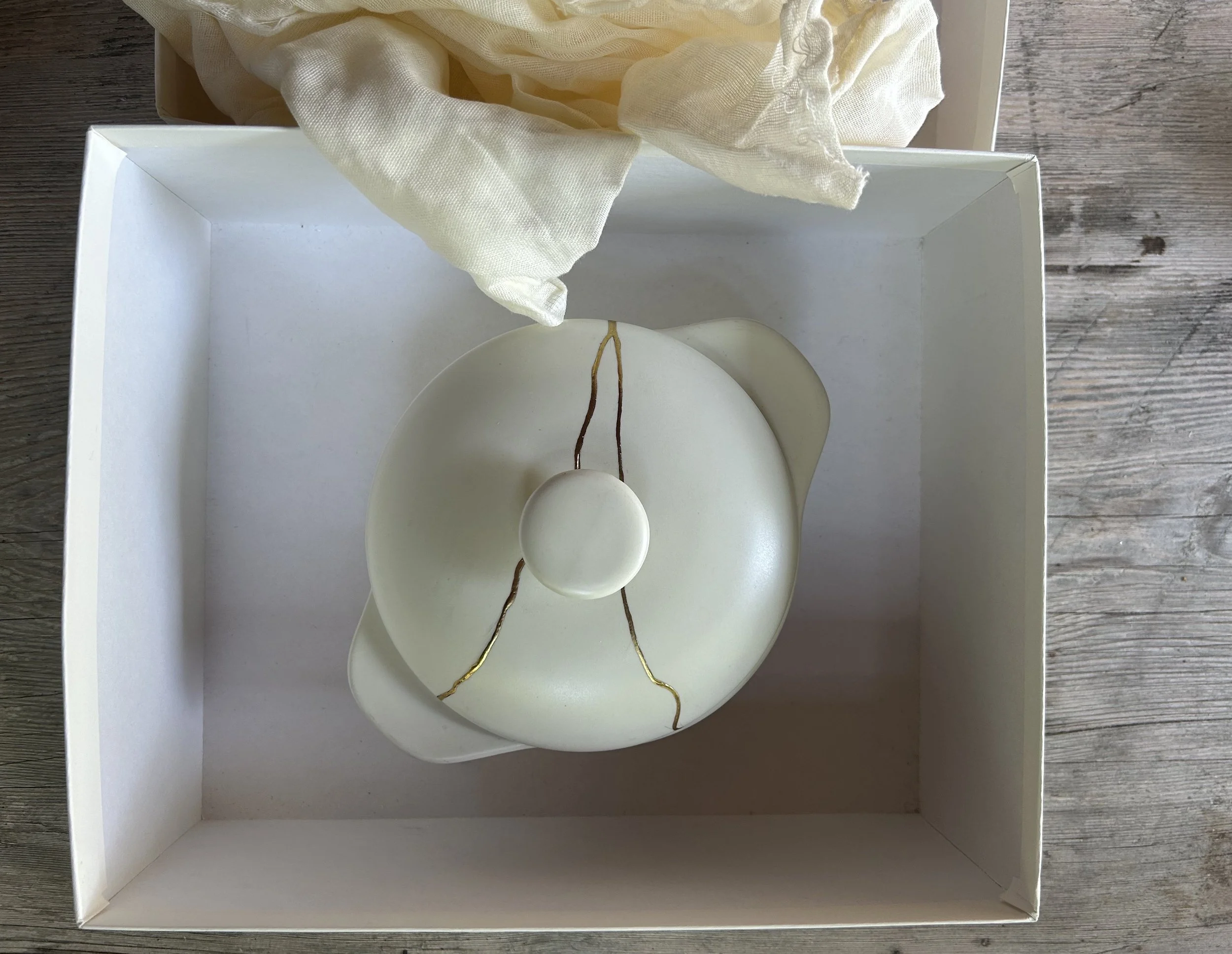 White ceramic dish with lid sitting in gift box, golden Kintsugi repair across surface