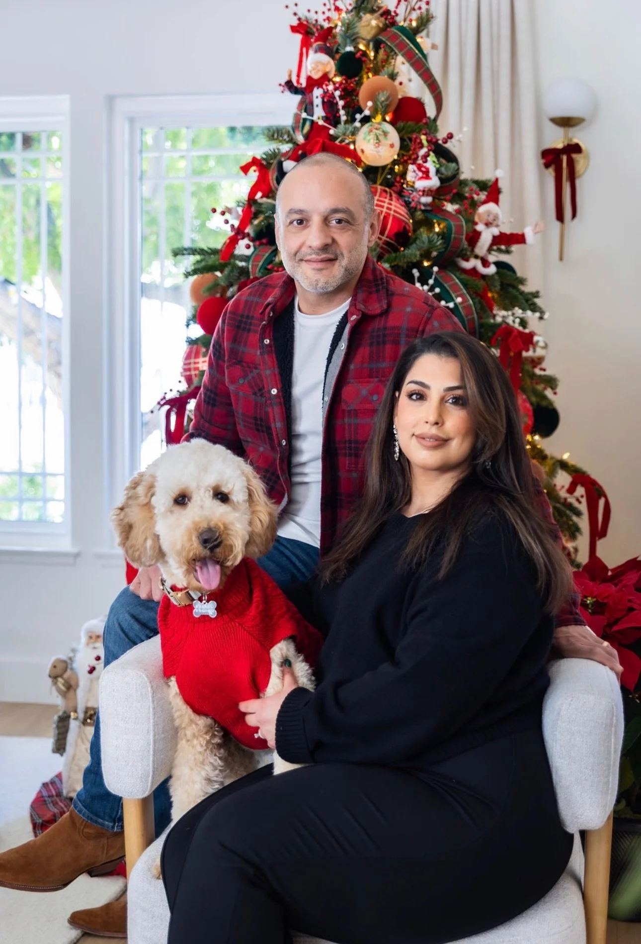 Goldendoodle family Christmas portrait with a couple seated by a decorated holiday tree indoors, featuring a curly cream Goldendoodle wearing a red sweater in a warm, cozy home setting