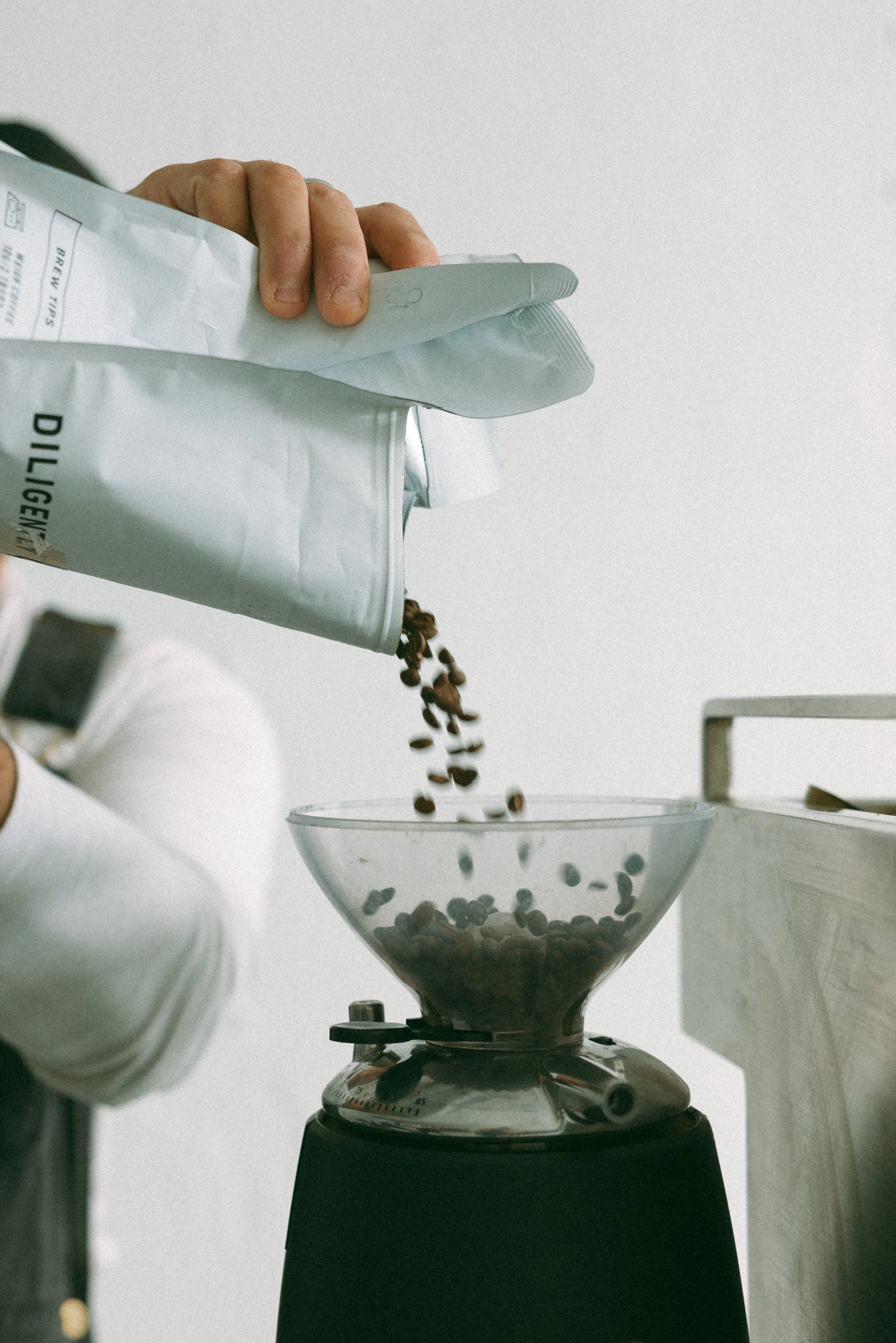 Hand pouring coffee beans from a bag into a glass coffee grinder.