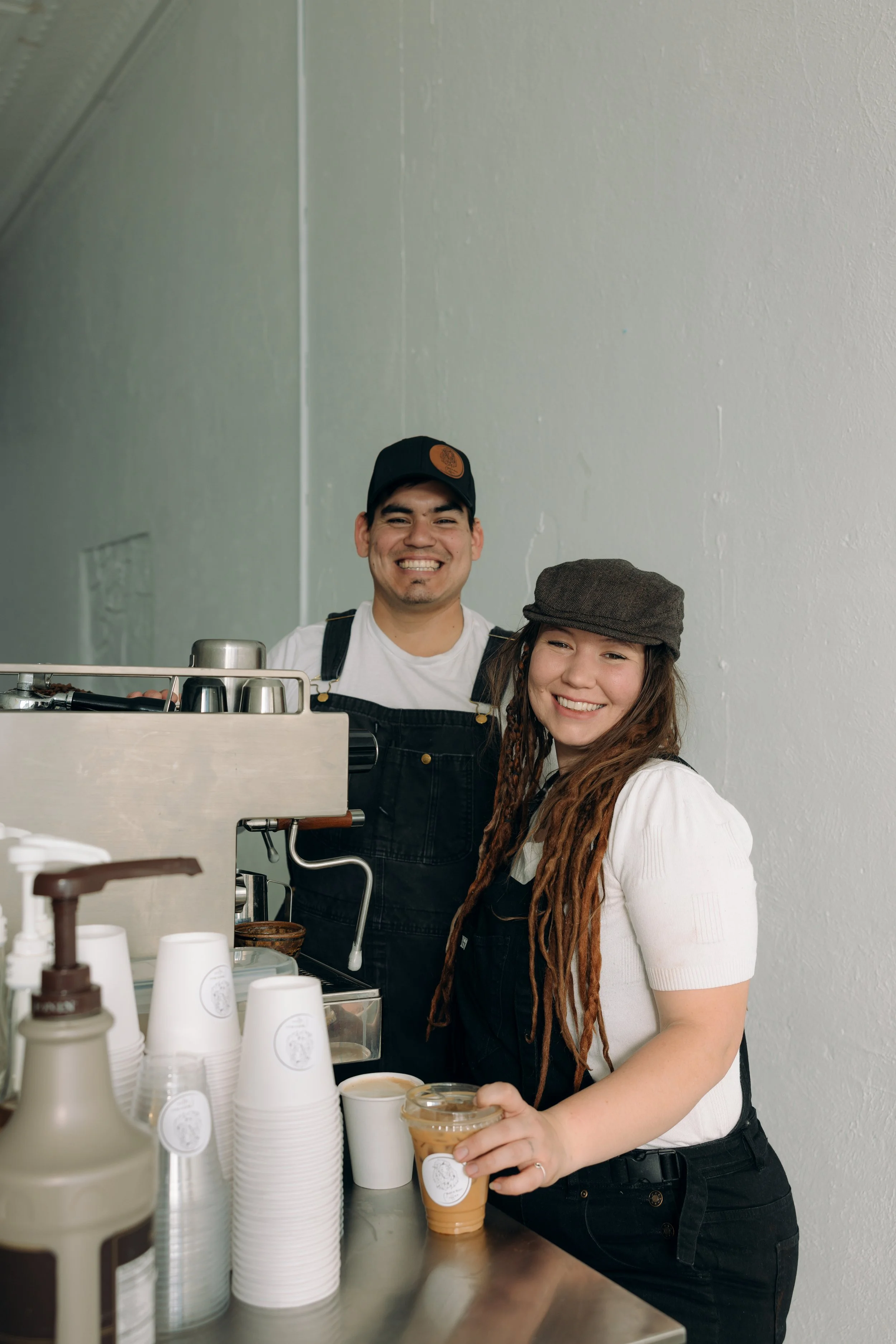 A smiling man and woman working behind a coffee shop counter with coffee cups and coffee-making equipment. At a coffee truck.