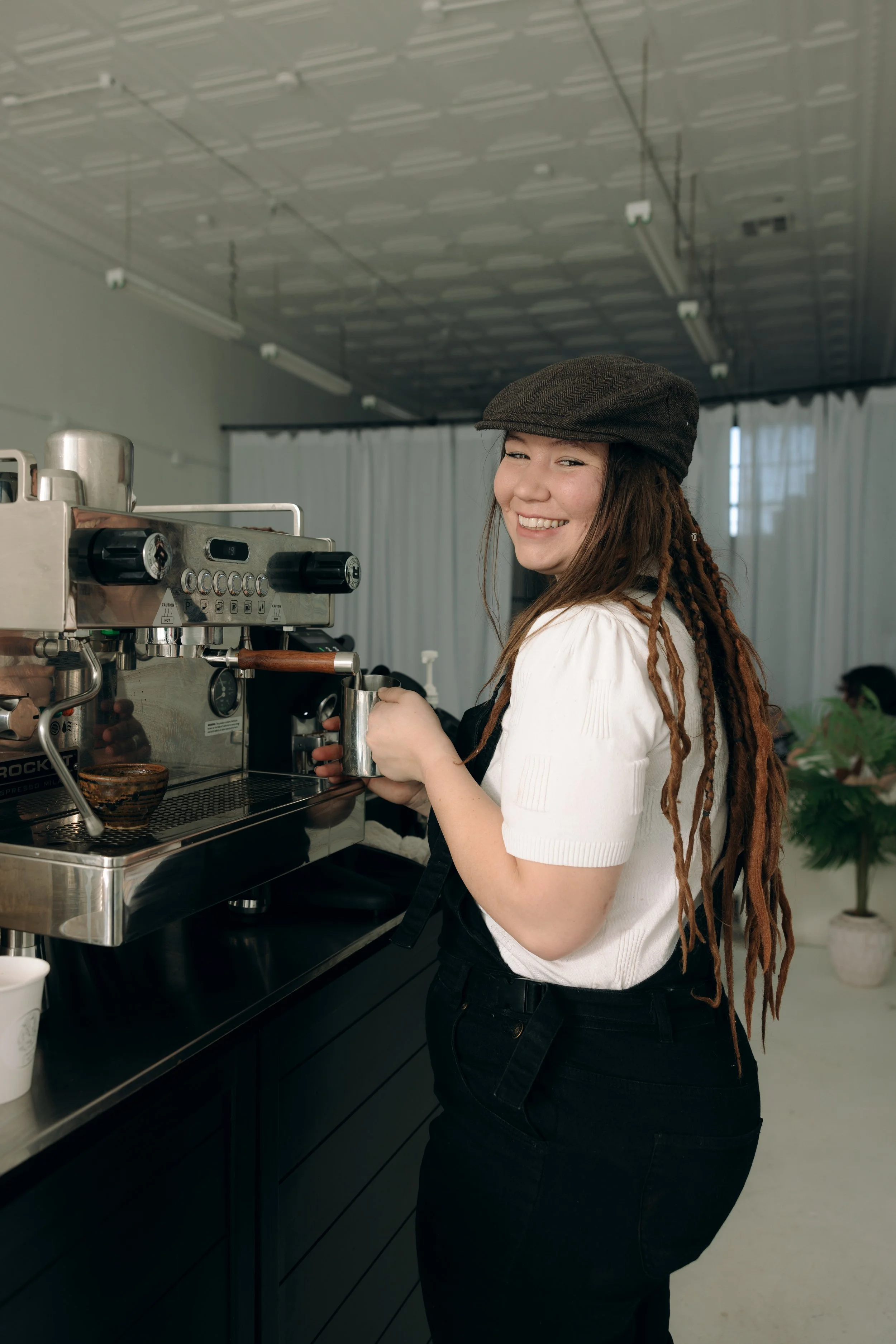 A woman with long dreadlocks, wearing a brown cap and white shirt, smiling while preparing coffee with an espresso machine in a modern cafe.