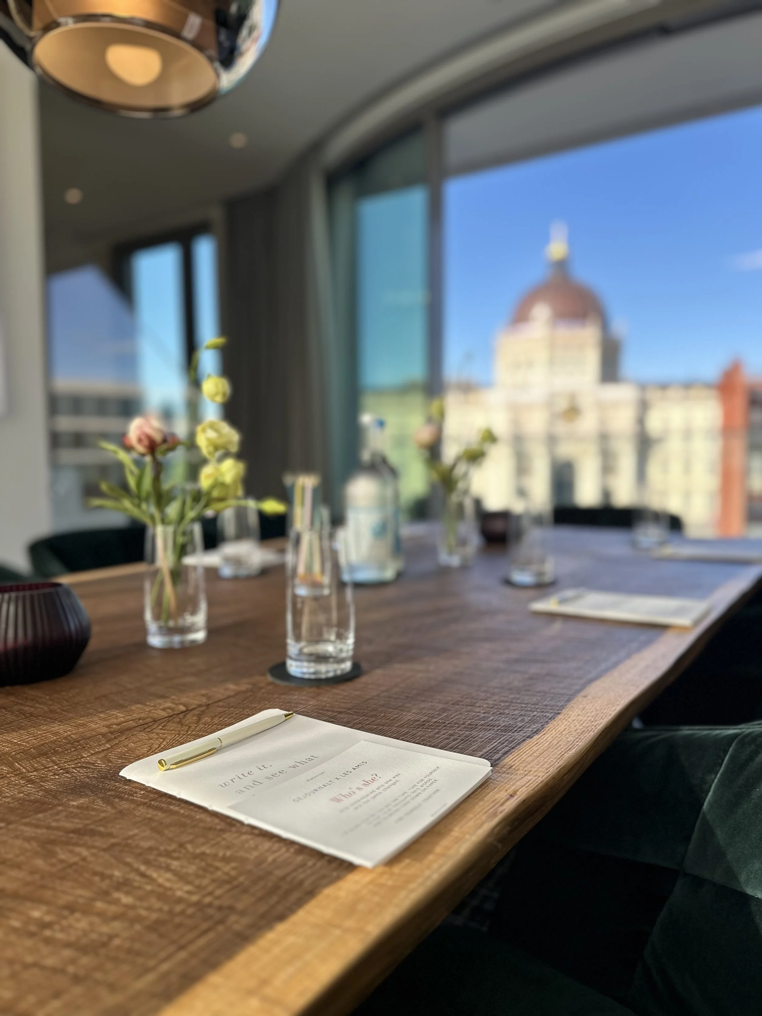 Ein Tisch in einem Restaurant mit Dekoration, Wasserflaschen, Gläsern und einer Karte, vor großen Fenstern mit Blick auf ein historisches Gebäude mit Kuppel und blauen Himmel.