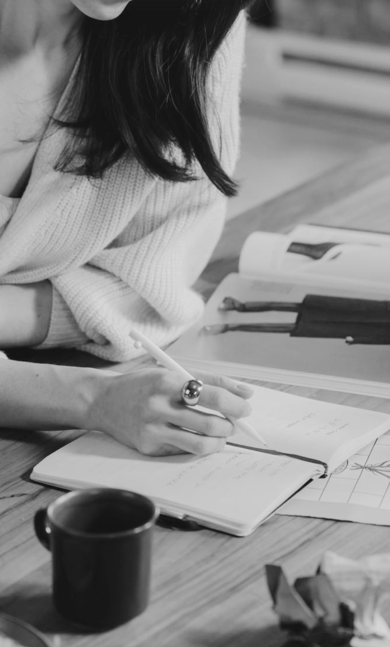Eine Person schreibt in einem Notizbuch, während sie auf einem Tisch mit verschiedenen Zeitschriften, einer Tasse und Kaffeetassen sitzt.
