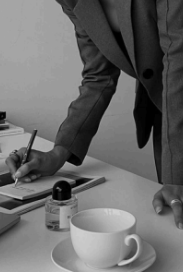 Geschäftsmann schreibt in einem Notizbuch auf einem Schreibtisch, auf dem auch eine Tasse, ein Flasche Teelöffel und einige andere Gegenstände stehen.