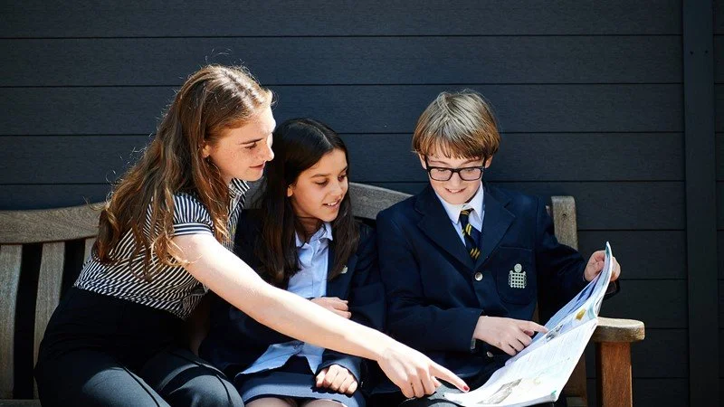 Girl and boy 11+ with their teacher sat on a bench in the sunshine in the playground of a London co-ed school.
