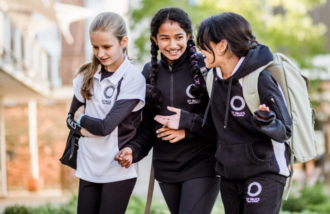 Three pupils from a highly academic 11+ entry private girls' school in black and white sports kit walking together, smiling and talking. They are outdoors in a school setting with blurred buildings and greenery in the background.