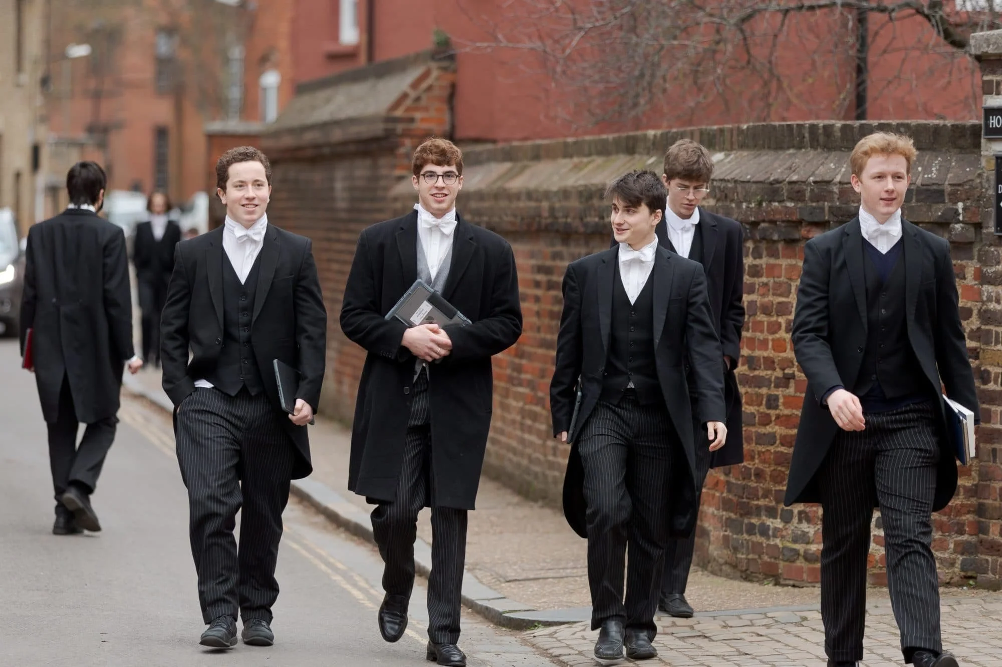 Group of young men in formal attire, including tailcoats and white bow ties, walking on a street with brick buildings in the background.